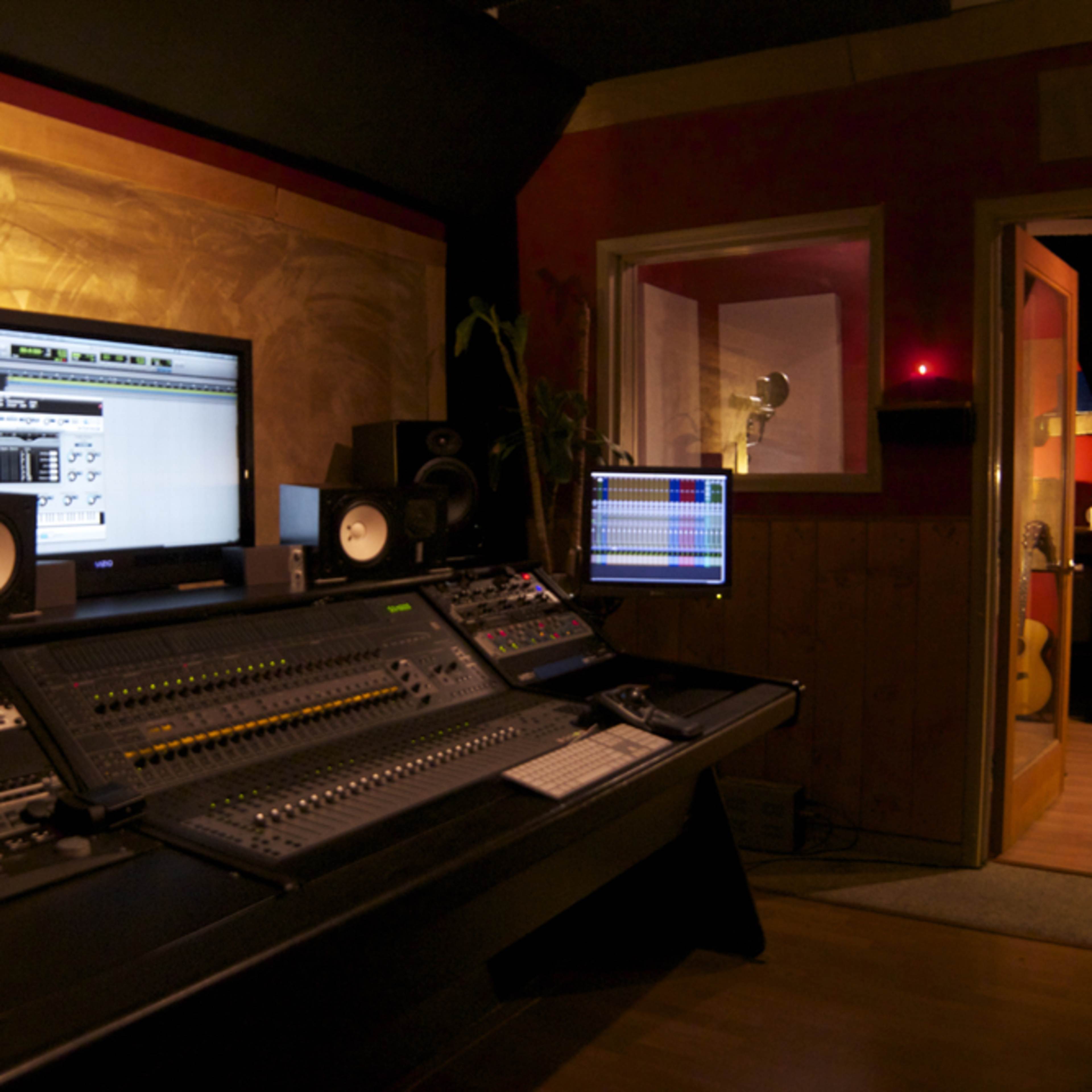 A professional music recording studio with a control console, computer monitors, and a hallway leading to a room with guitars.