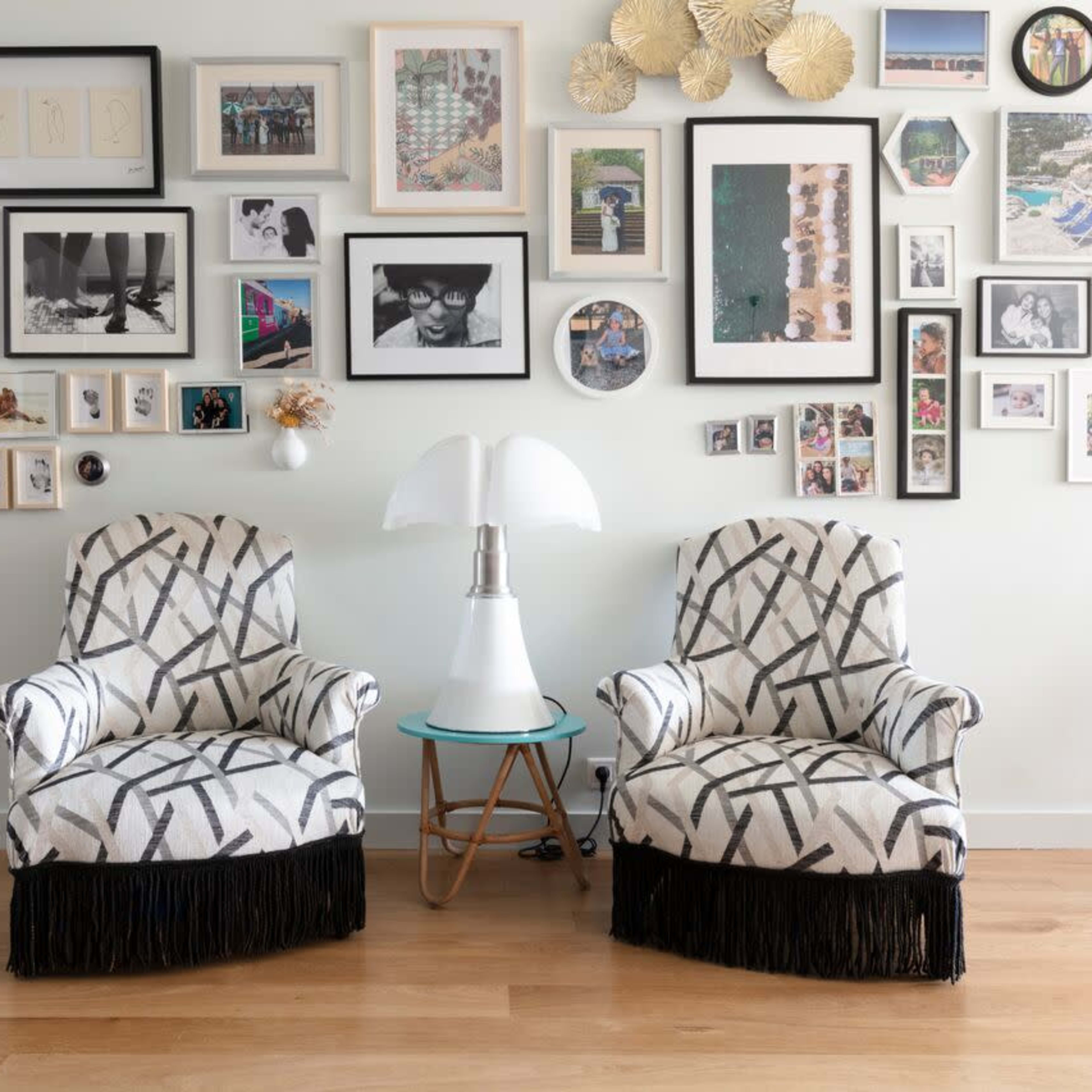 Two patterned armchairs with fringe placed in front of a gallery wall filled with framed photographs and artwork, alongside a small lamp and table.