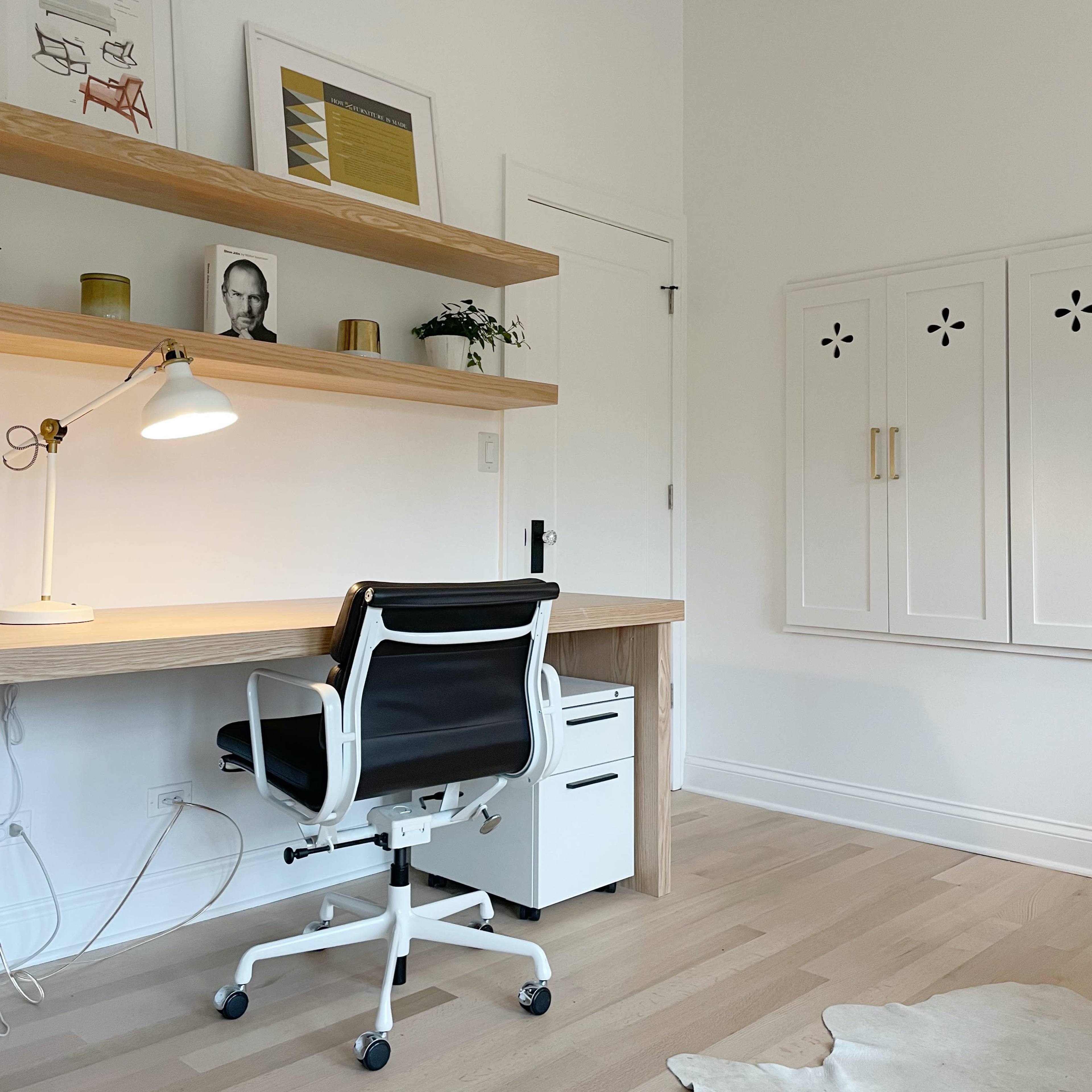 A minimalist home office features a wooden desk with a chair, a lamp, and shelves displaying framed art and a potted plant.