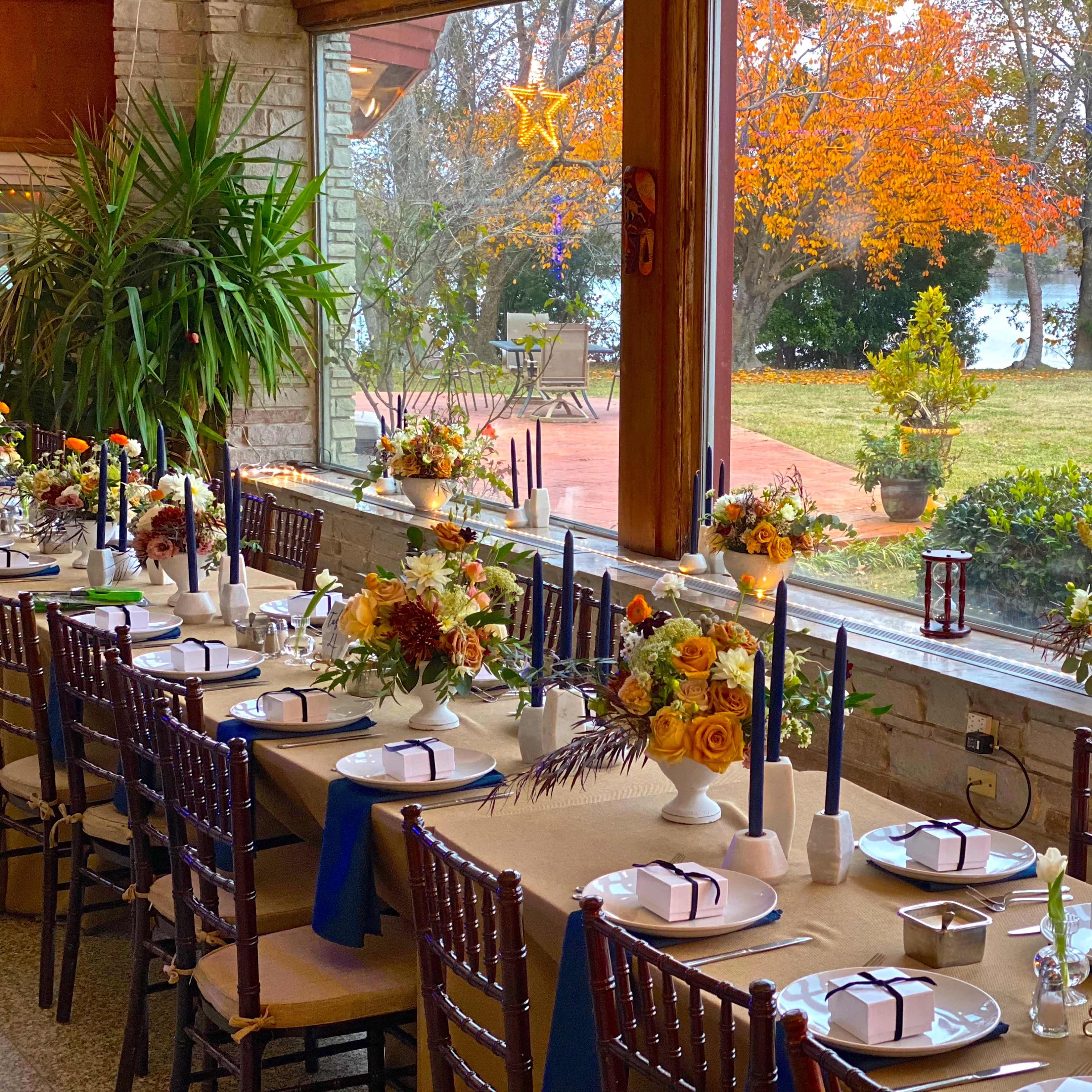 A long dining table is set with floral centerpieces and candles, overlooking a lake with autumn trees in the background.