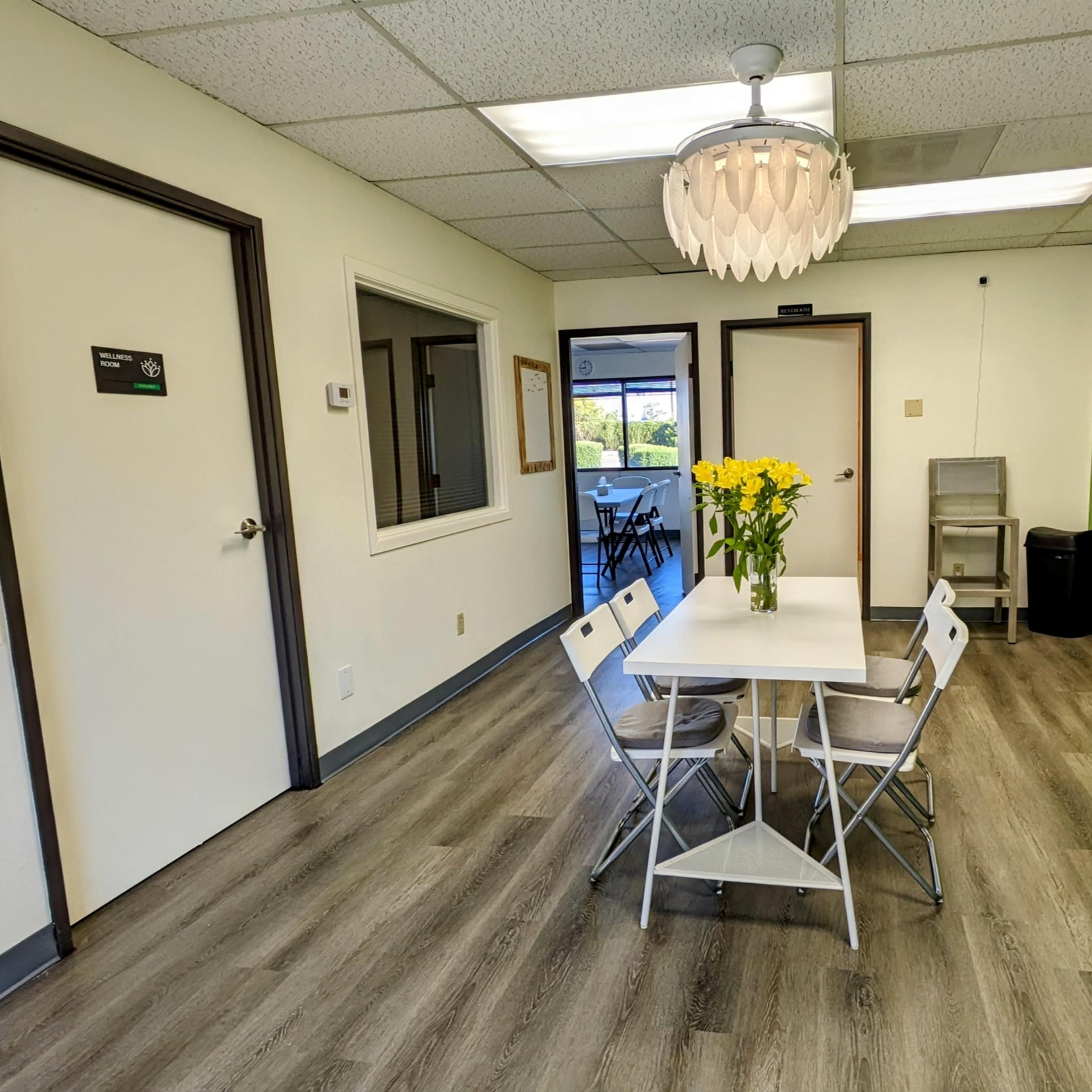 A bright, modern waiting room features a white table surrounded by four folding chairs, green accent walls, and potted plants.
