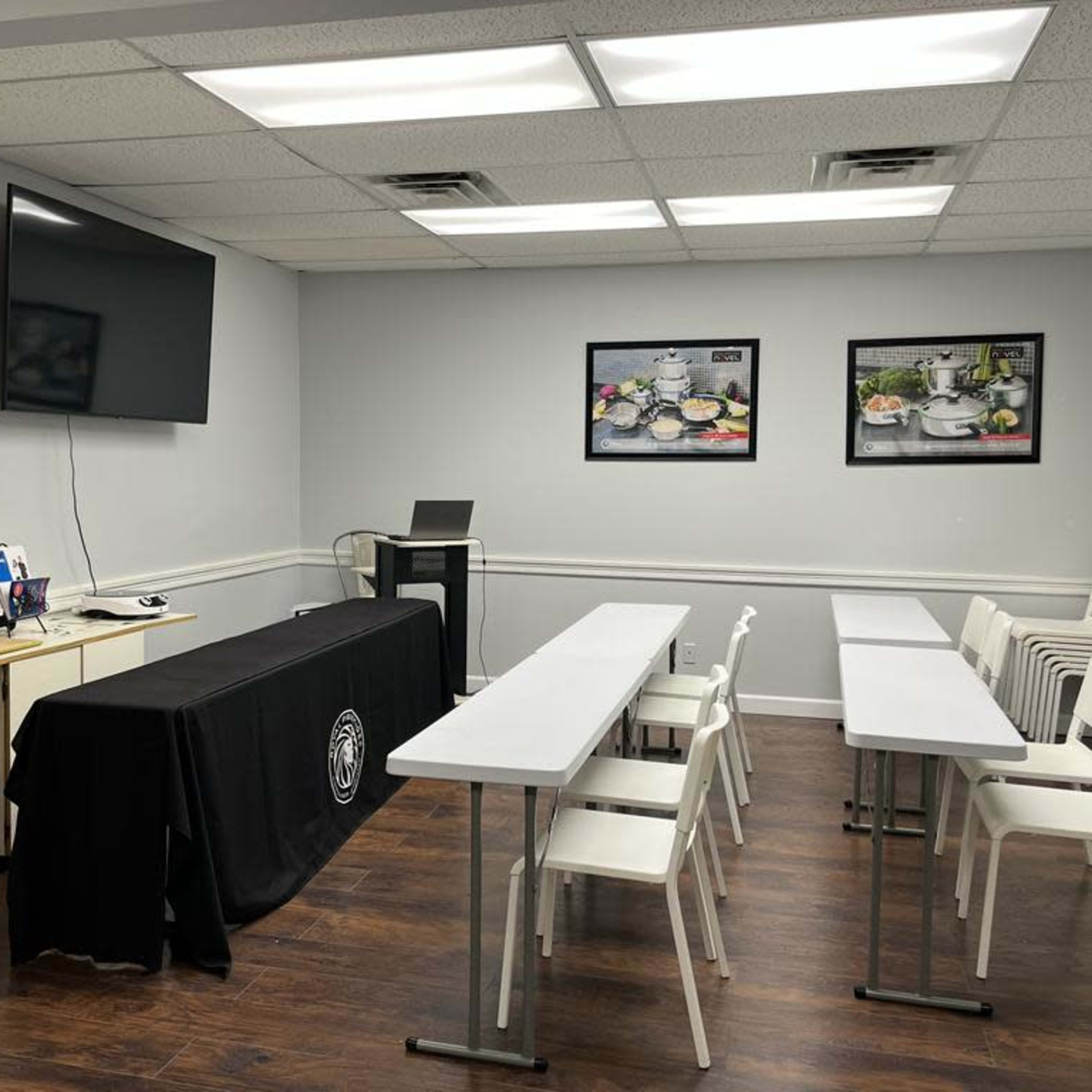 A classroom setting with a long table covered by a black tablecloth, several white chairs arranged around tables, and a large screen mounted on the wall.