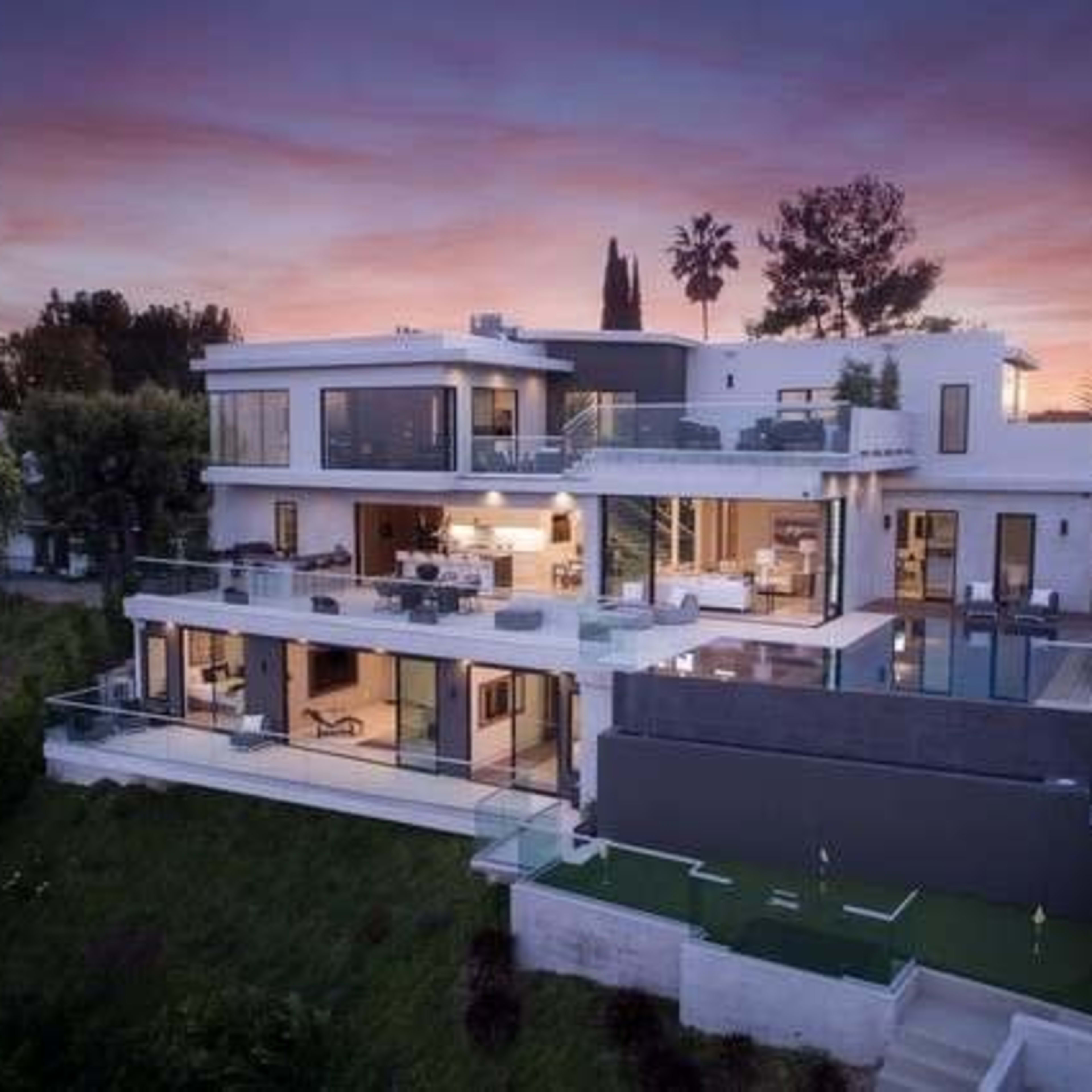 A modern multi-level house with large glass windows and a terrace, situated on a hillside at sunset.
