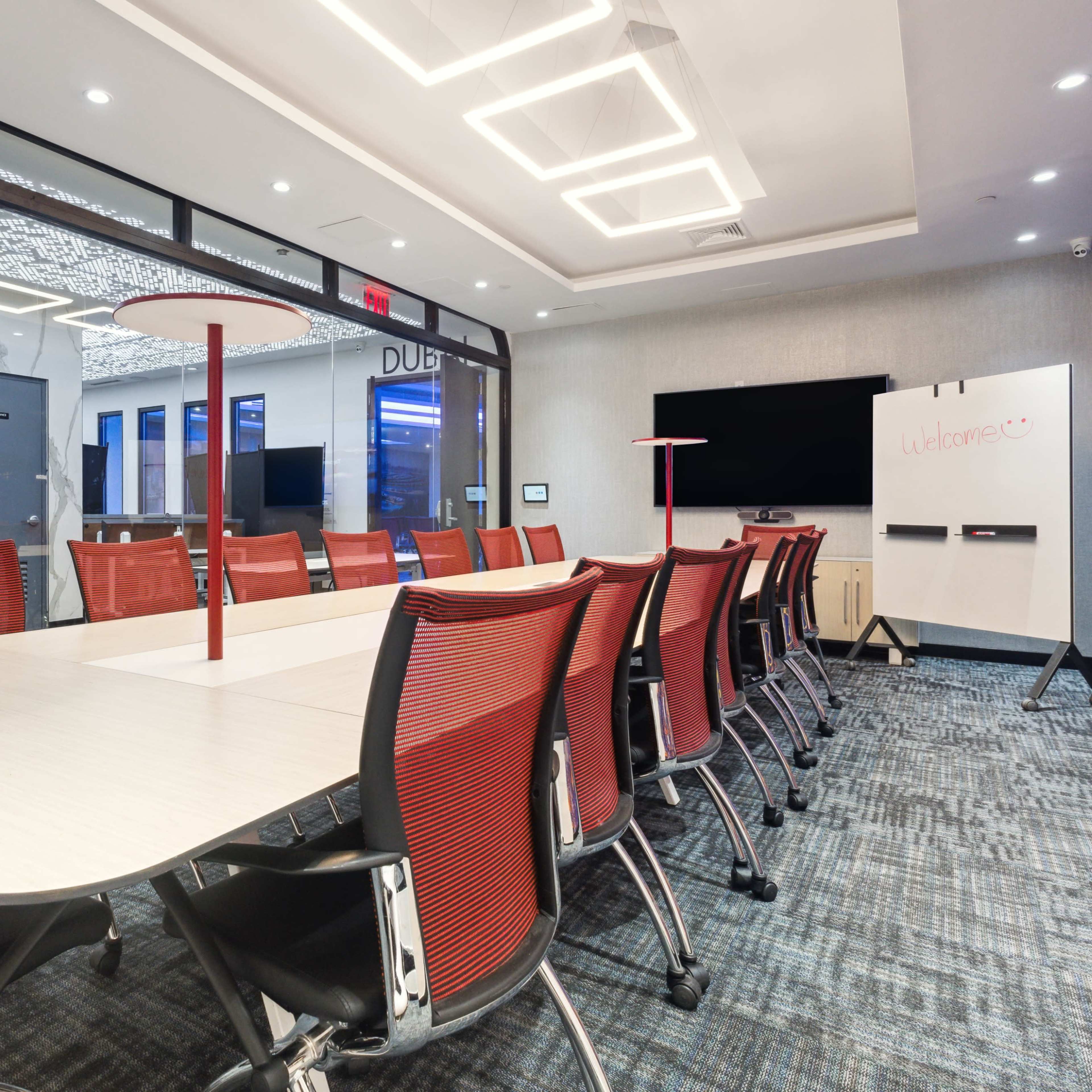 A modern conference room features a long table surrounded by red chairs, with a TV screen and whiteboard on opposing walls.