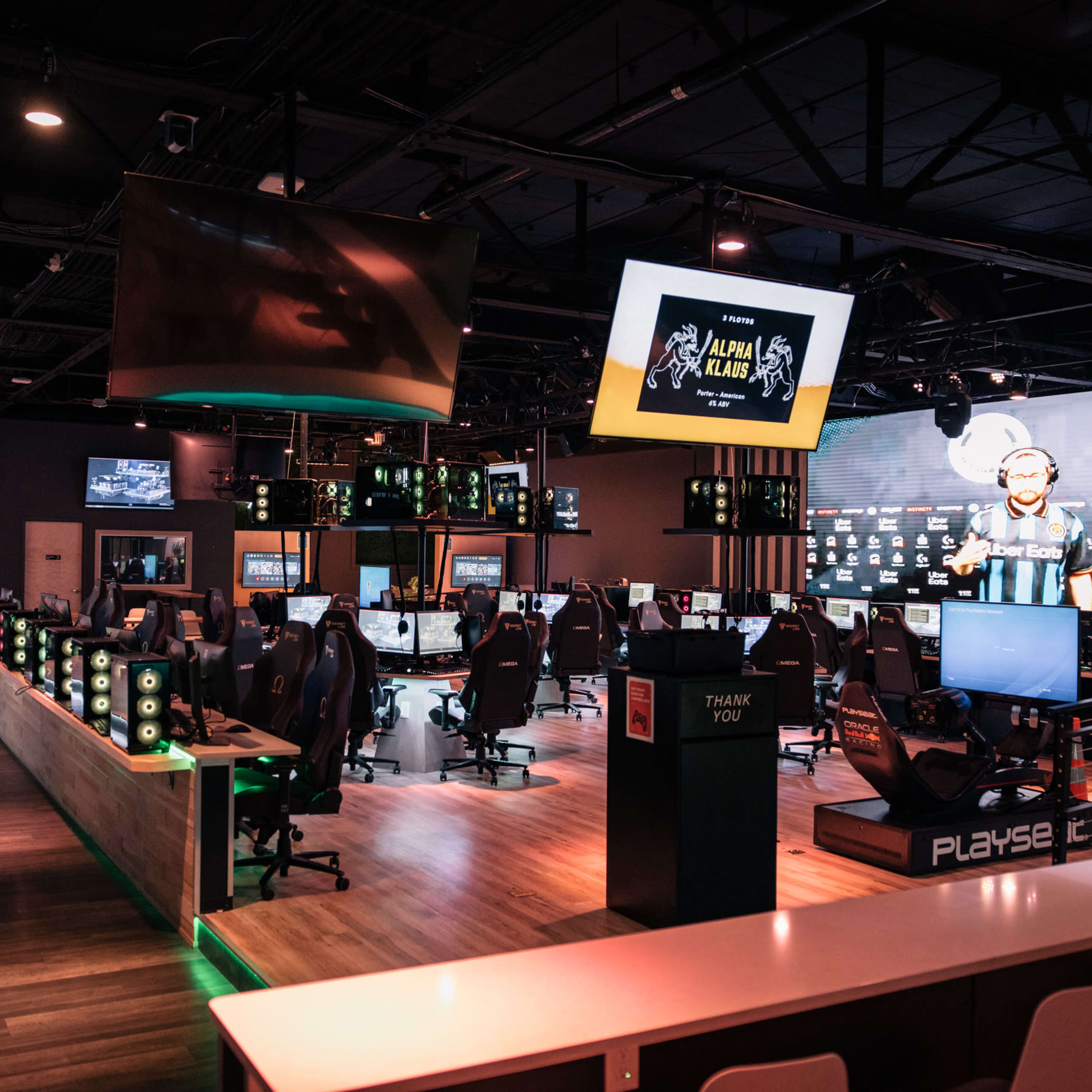 A modern esports gaming lounge features rows of high-end gaming computers, large screens displaying a tournament, and illuminated gaming accessories.