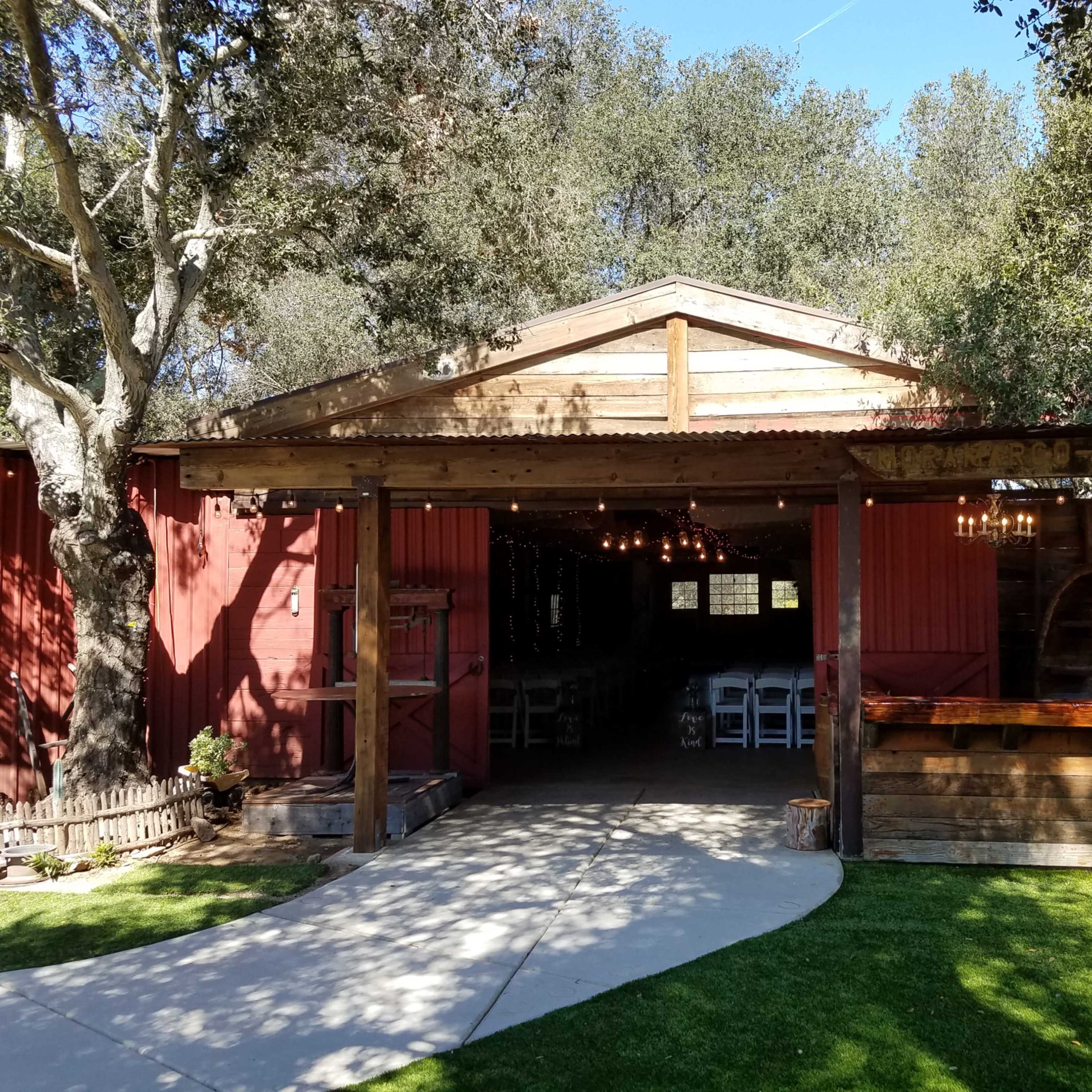 A rustic wooden structure with a corrugated metal roof features an open entrance, surrounded by trees and a manicured lawn.