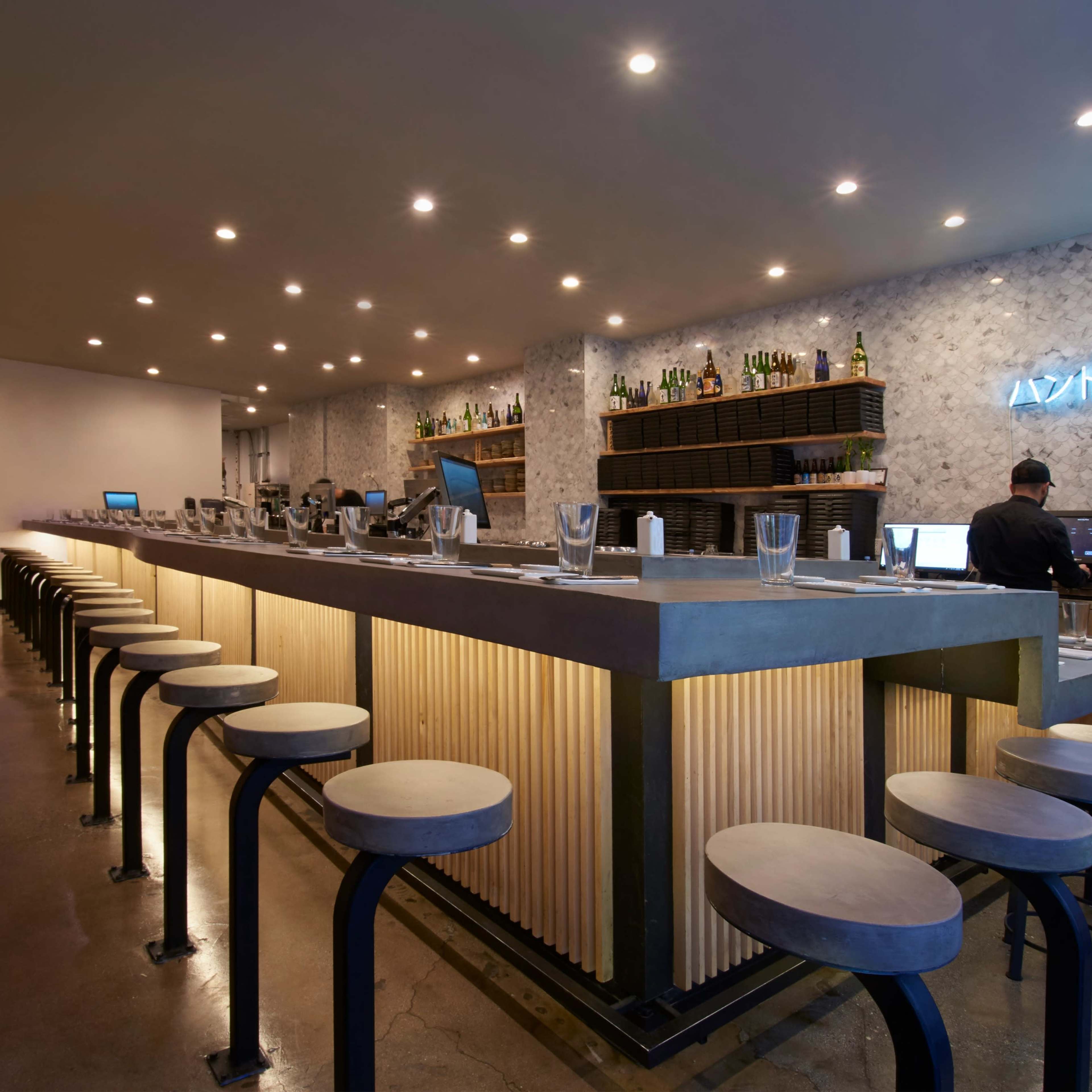 A modern bar interior with a long concrete counter, high stools, and warm lighting, complemented by a wall of bottles in the background.