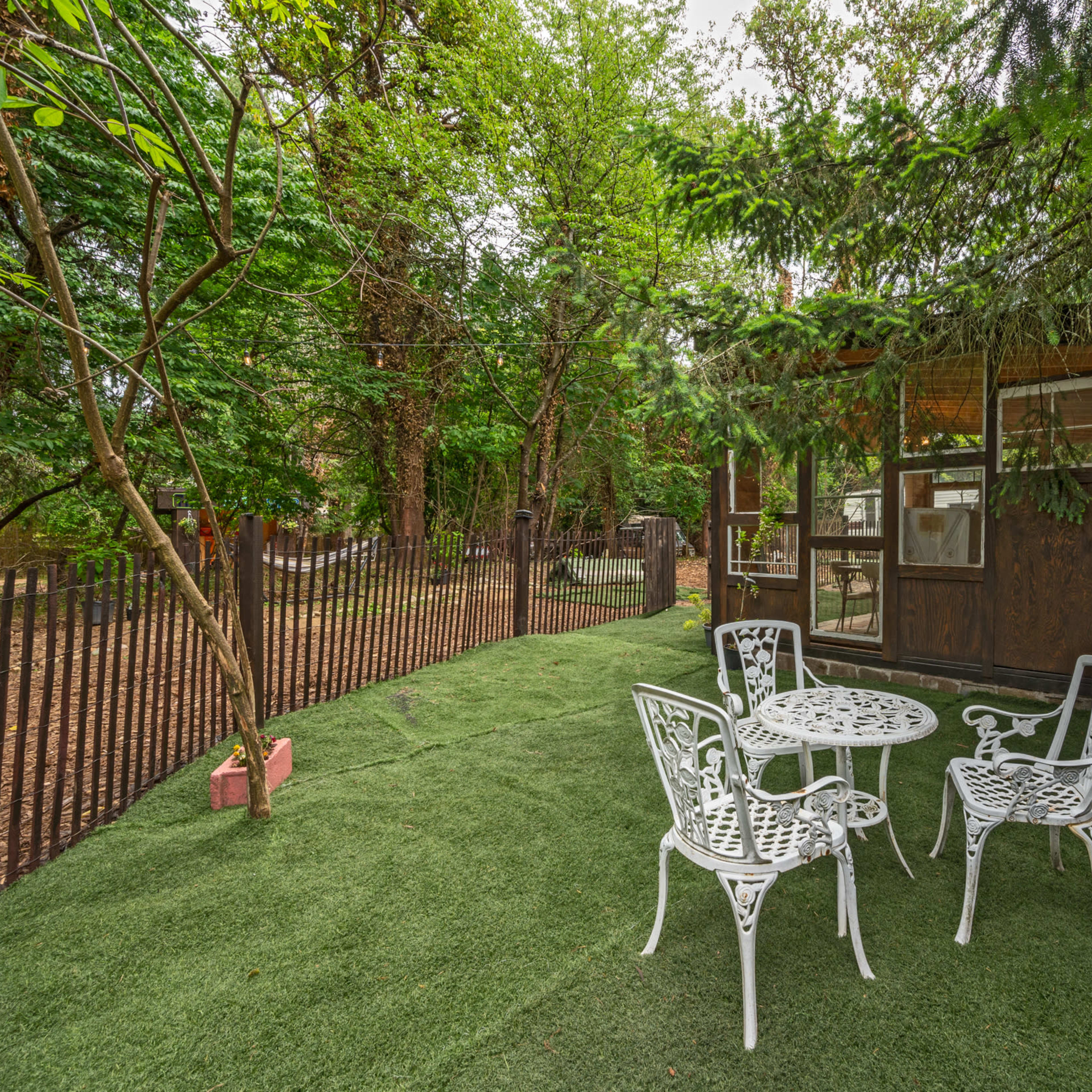 A small outdoor seating area with white metal furniture is set on artificial grass near a wooden cabin surrounded by trees.