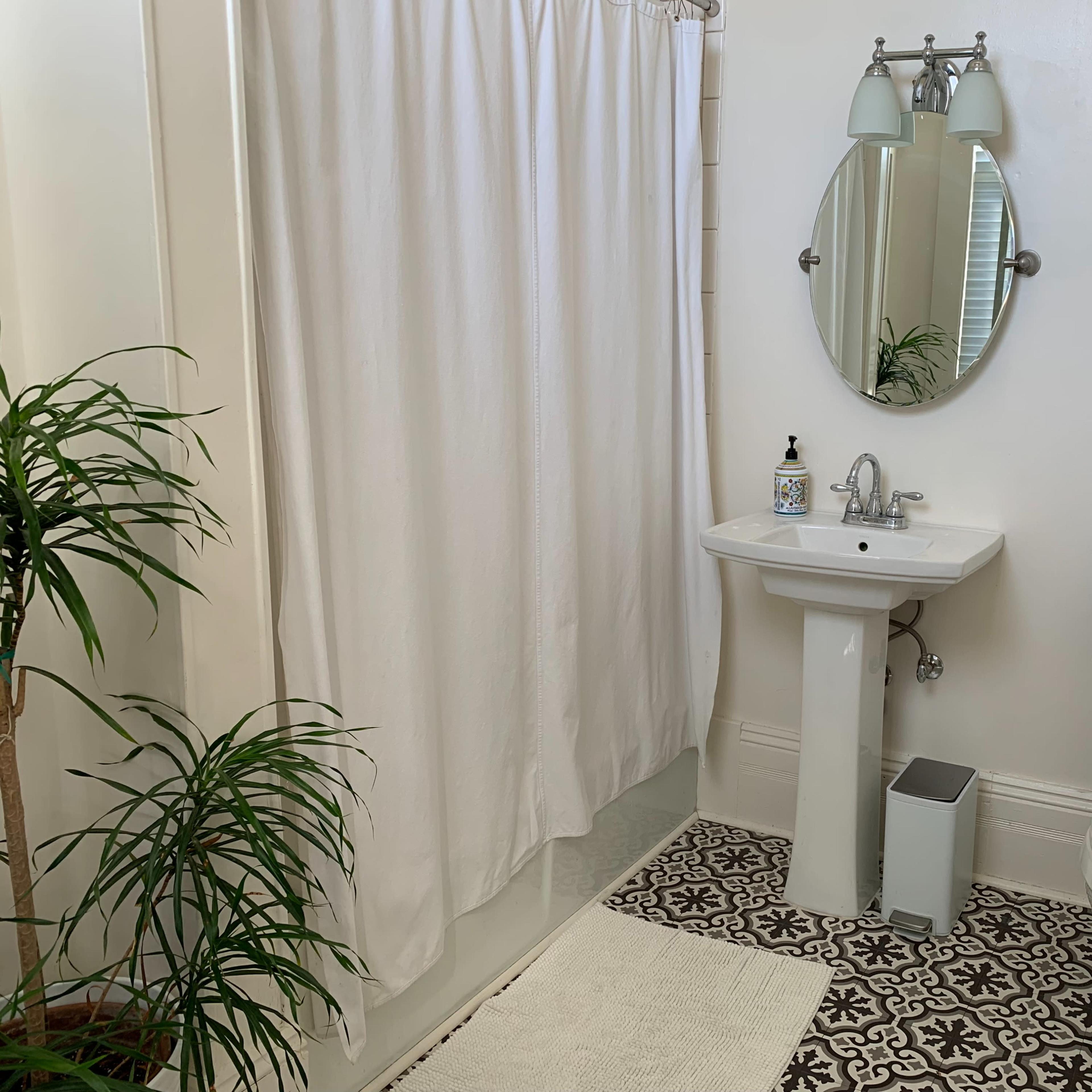 The bathroom features a white shower curtain, a round mirror above a pedestal sink, a toilet, and decorative floor tiles.