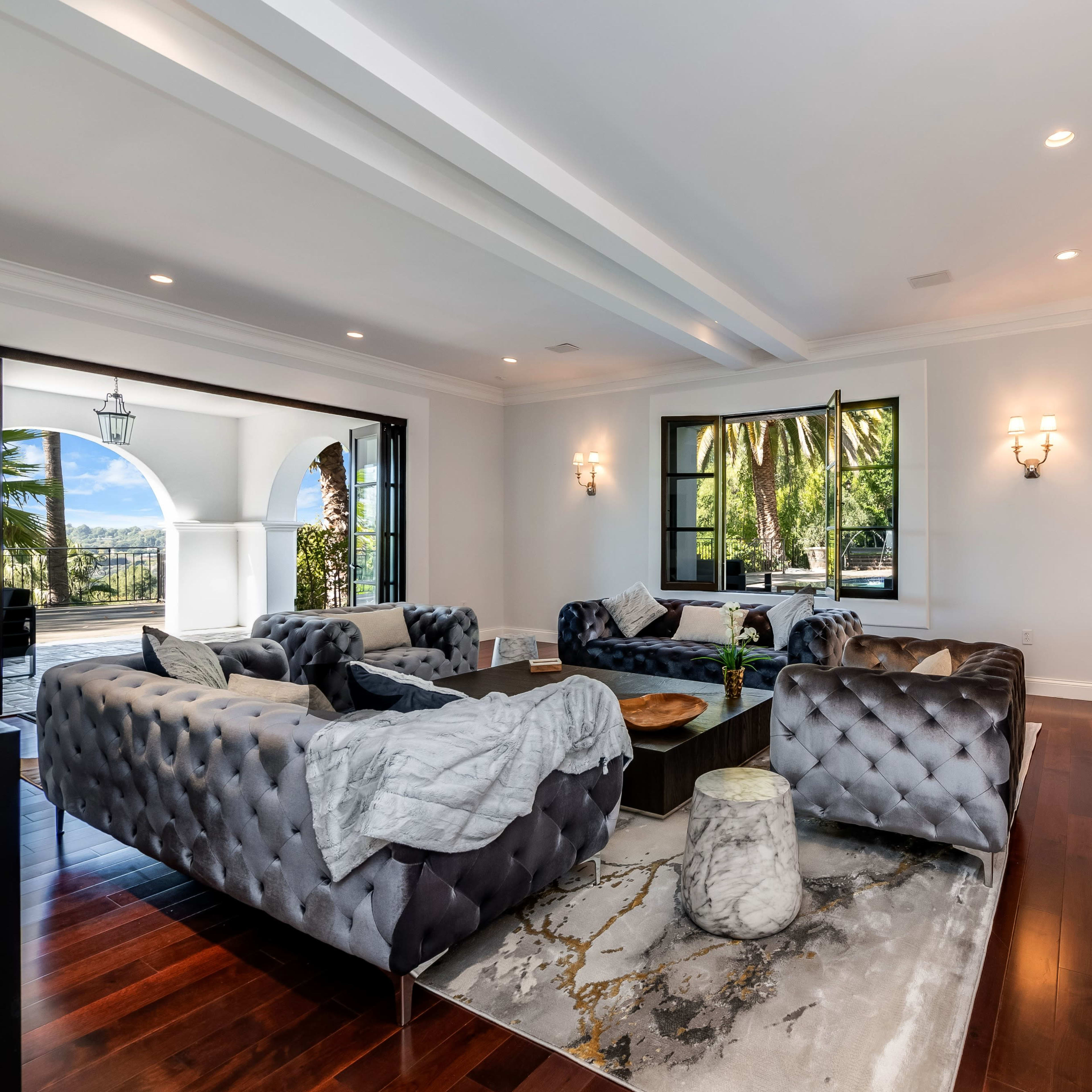A spacious living room with tufted velvet sofas, a marble fireplace, and large windows framing a view of palm trees.