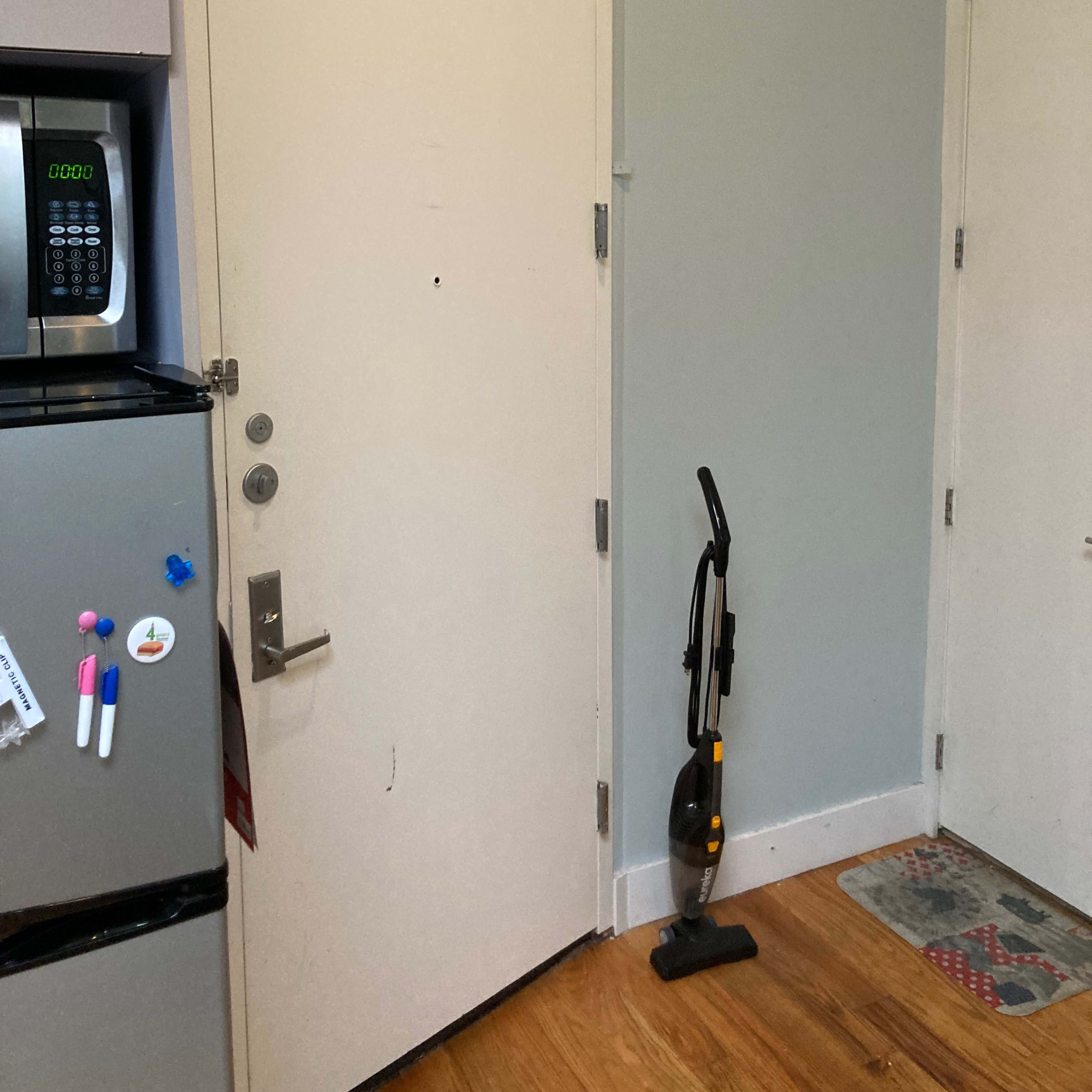 A vacuum cleaner stands upright against a wall near a closed door in a kitchen area with a refrigerator and a microwave.