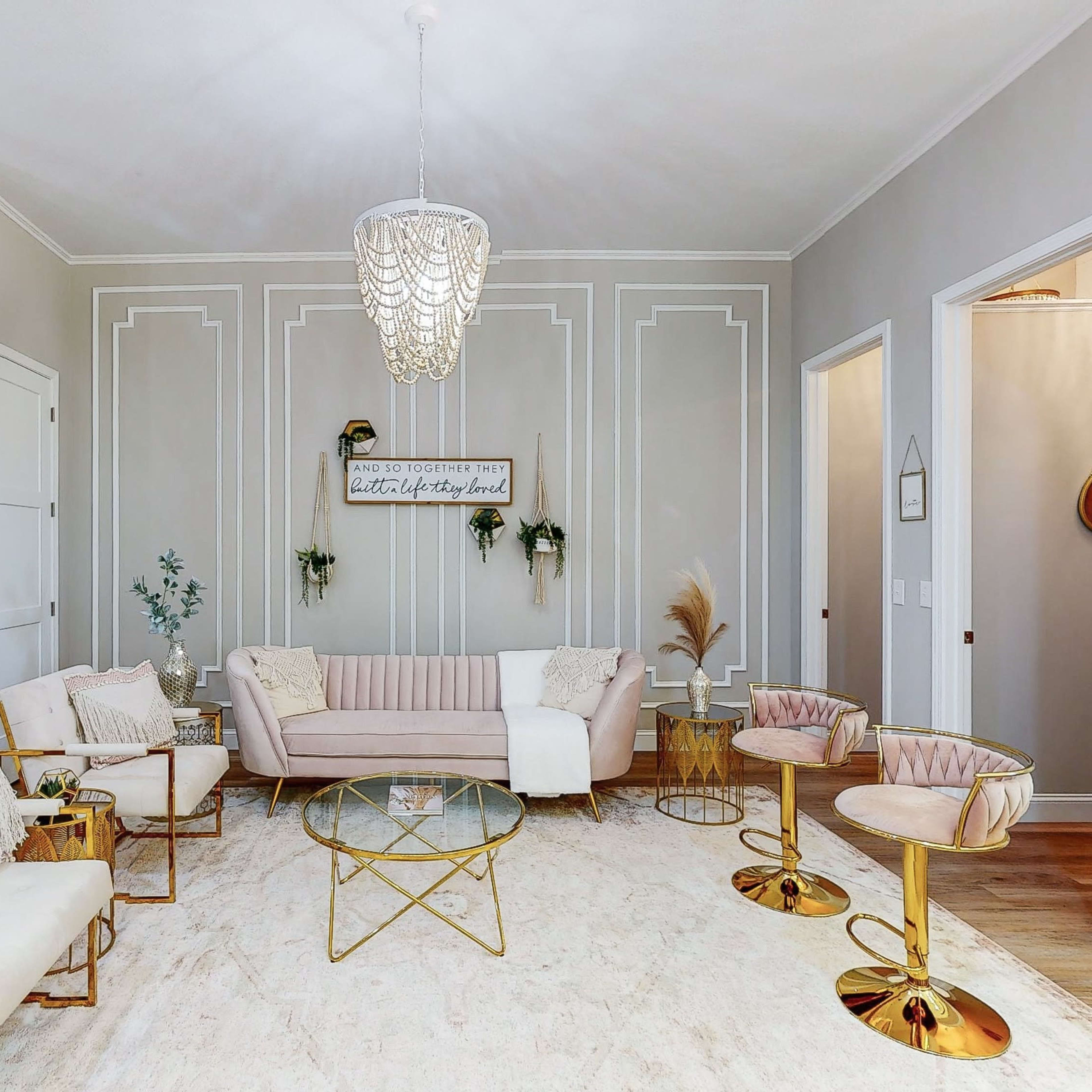 A modern living room features a light-colored sofa and armchairs arranged around a glass coffee table on a large area rug, with decorative wall accents and pendant lighting.