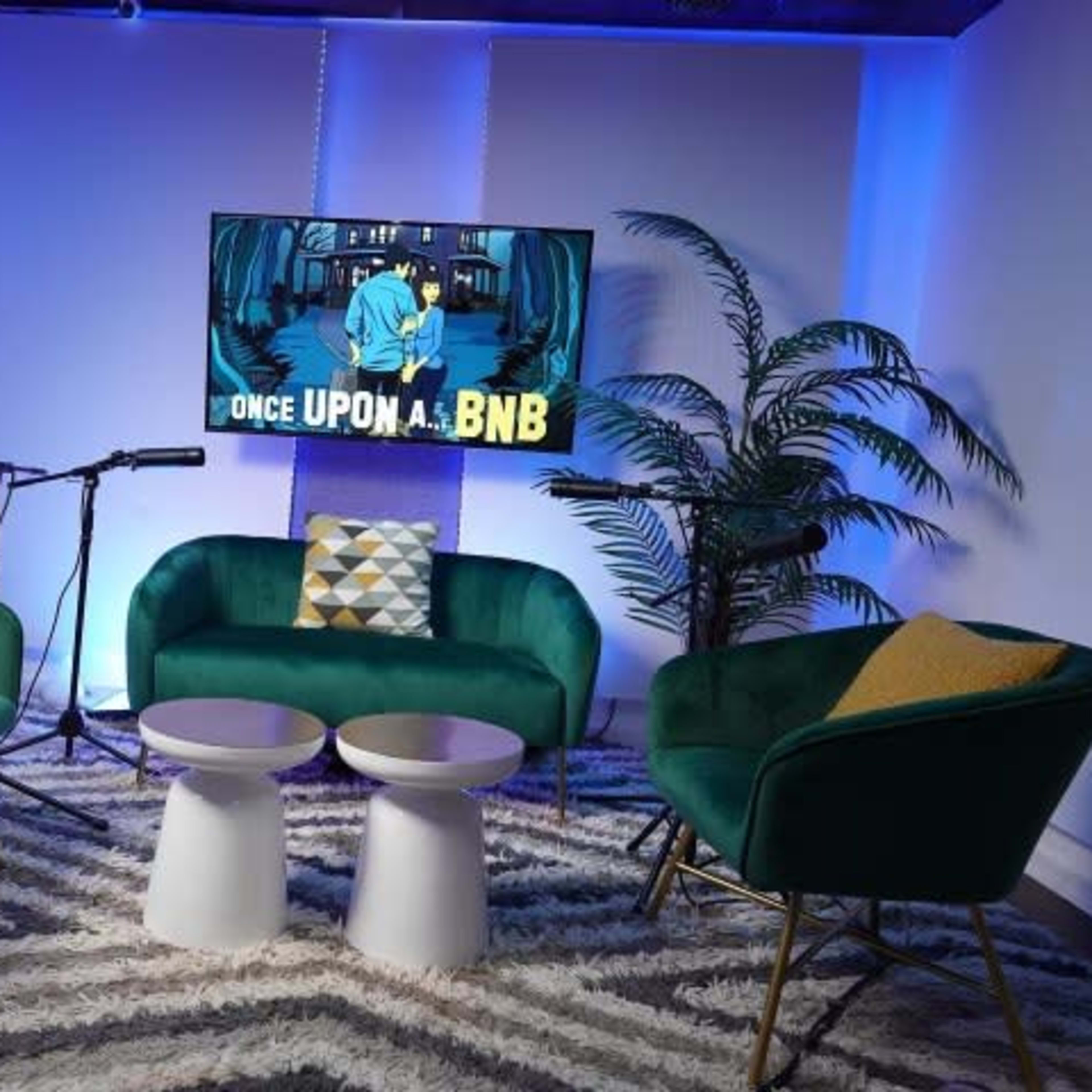 A modern living space features two green chairs and a teal sofa arranged around two white tables, with a large screen displaying "ONCE UPON A . . . BNB" set against a blue-lit backdrop.