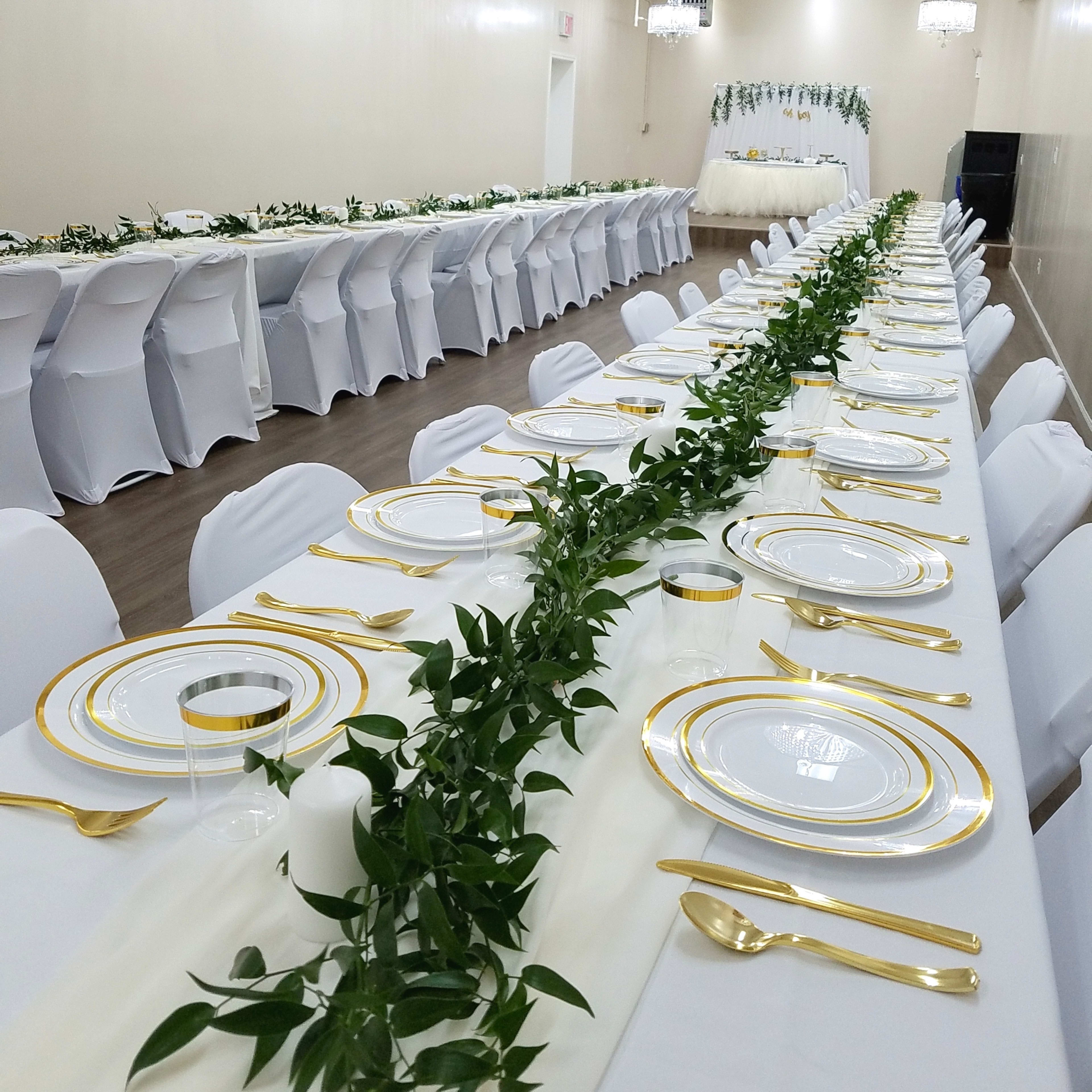 A long banquet table is set with white tablecloths, gold-rimmed plates, and greenery down the center, surrounded by white chairs in a well-lit hall.