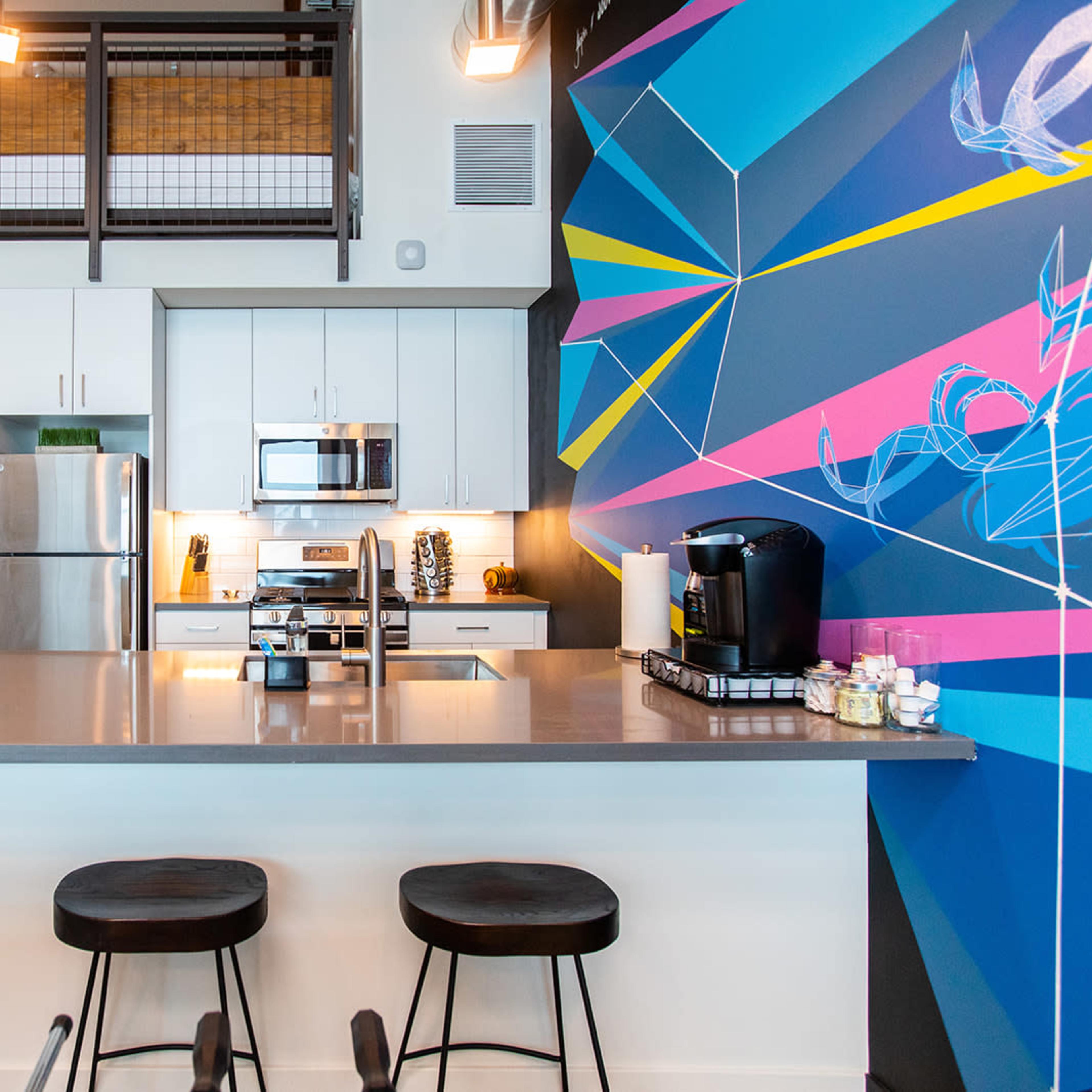 A modern kitchen with a colorful geometric wall mural, stainless steel appliances, and a sleek countertop with three black stools.