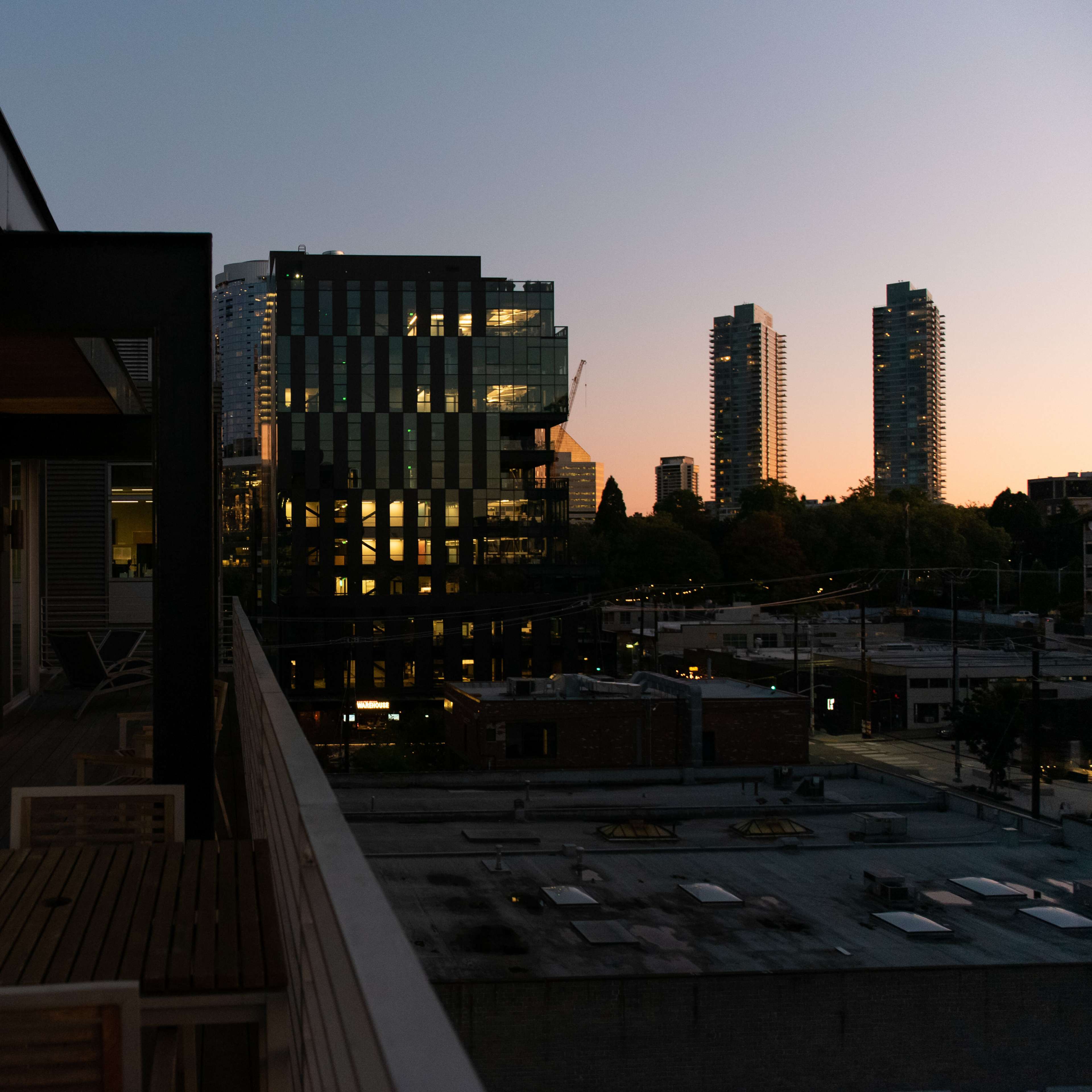 A balcony with wooden furniture overlooking a city skyline at dusk, with illuminated buildings and a colorful sunset in the background.