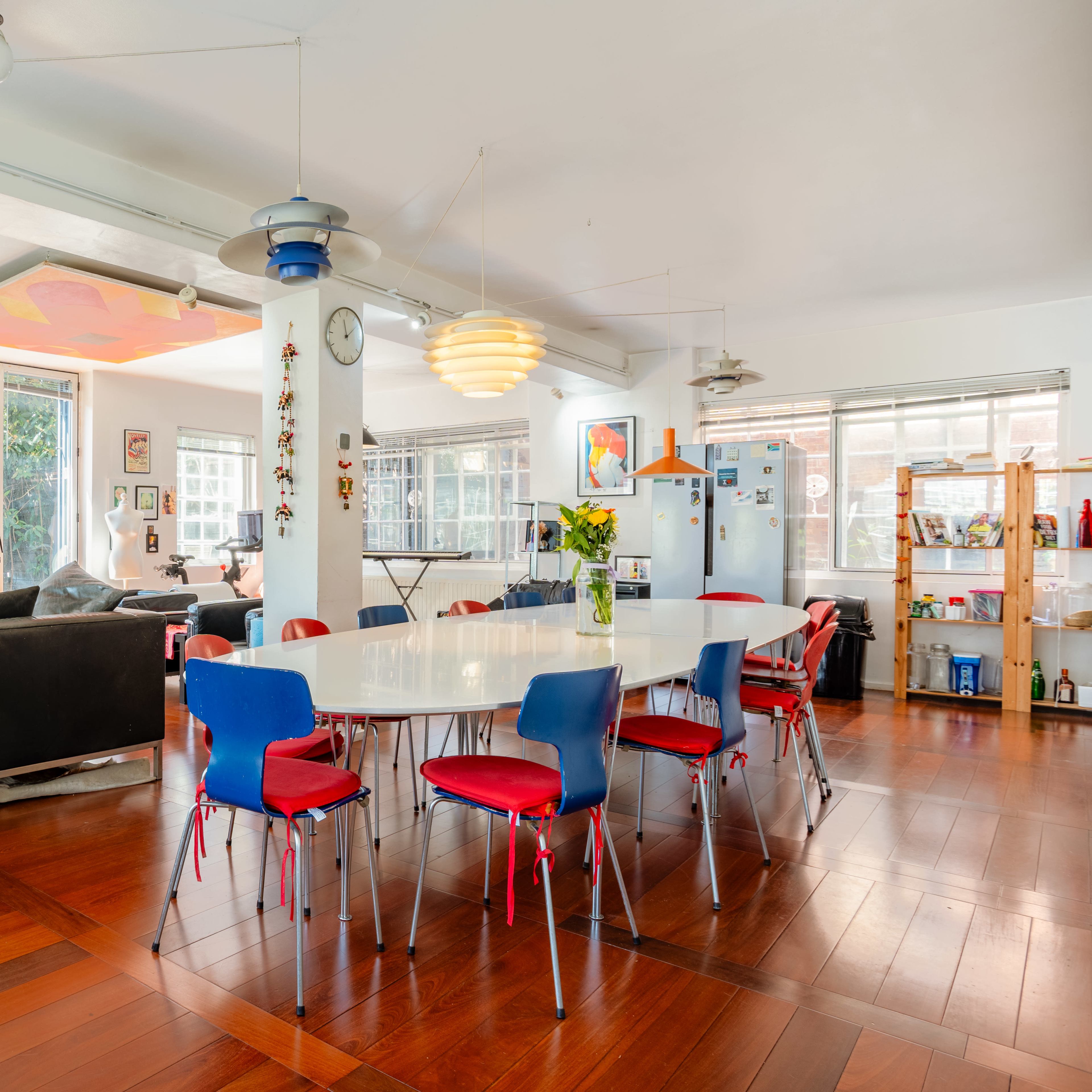 A spacious, modern open-concept living area features a dining table surrounded by colorful chairs, a large sofa, and multiple windows allowing natural light to fill the space.