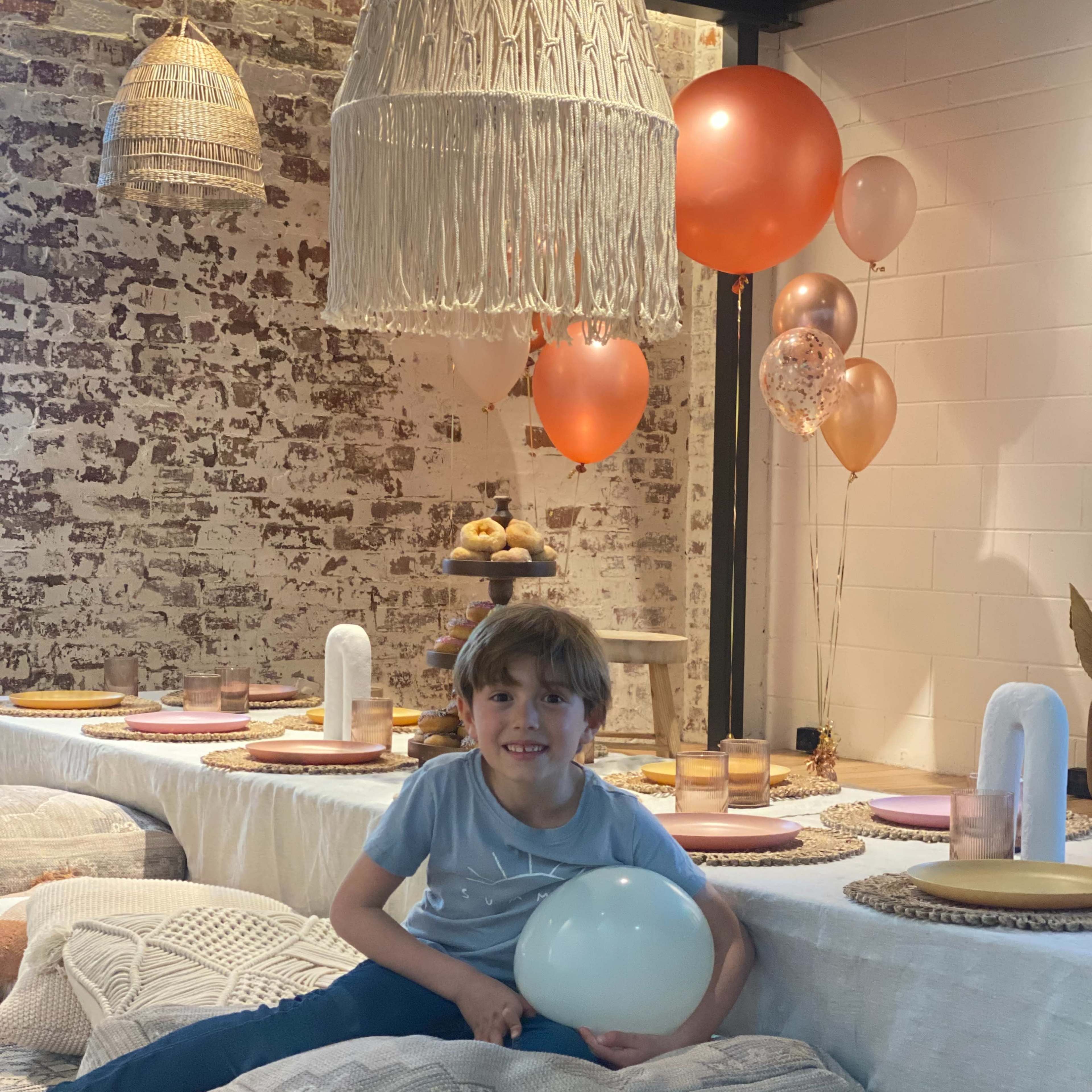 A young boy sits on a cushion in a decorated room with a festive table setup, balloons, and a variety of pastries.
