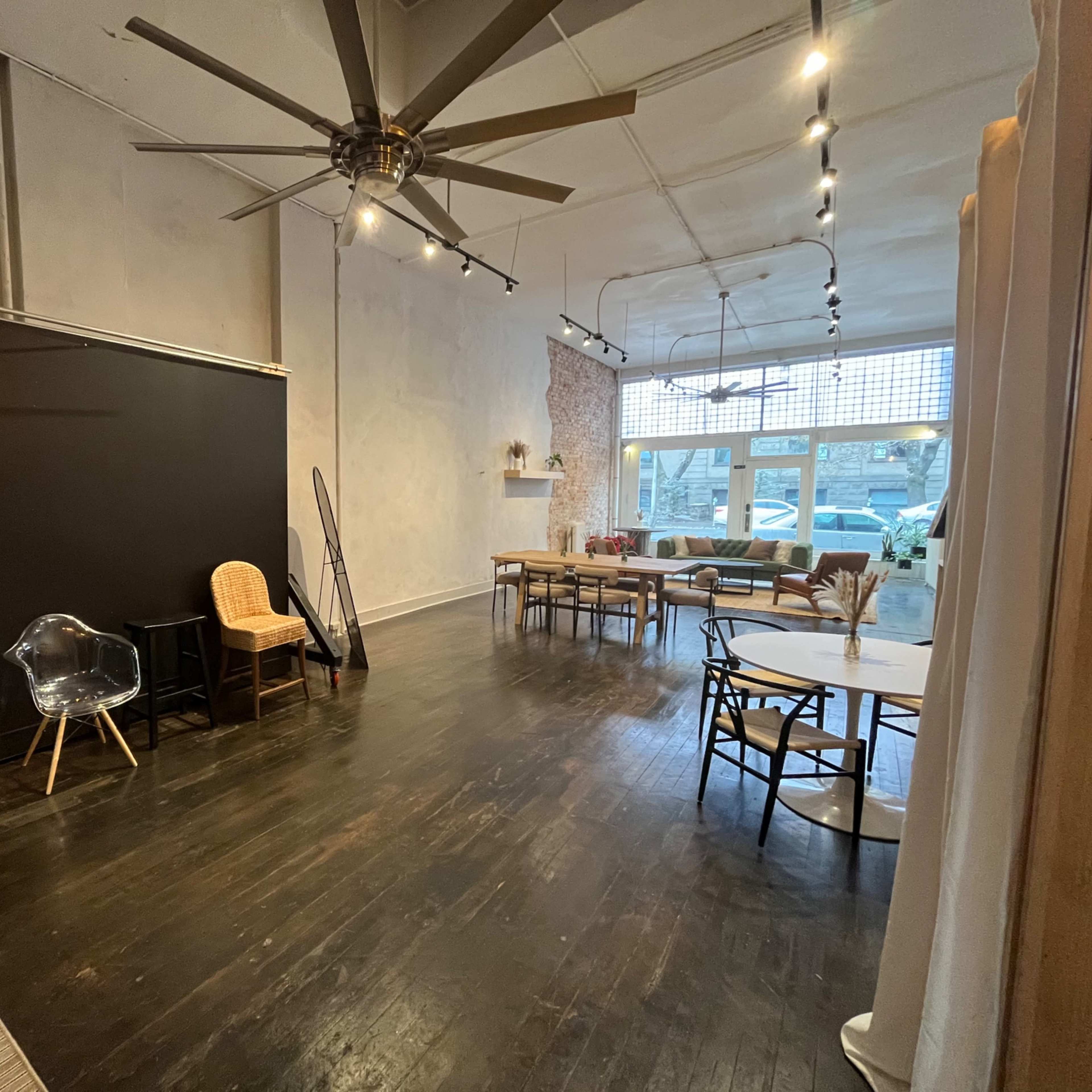 An empty interior space with a large ceiling fan, various seating options, a black backdrop, and a row of windows along one wall.