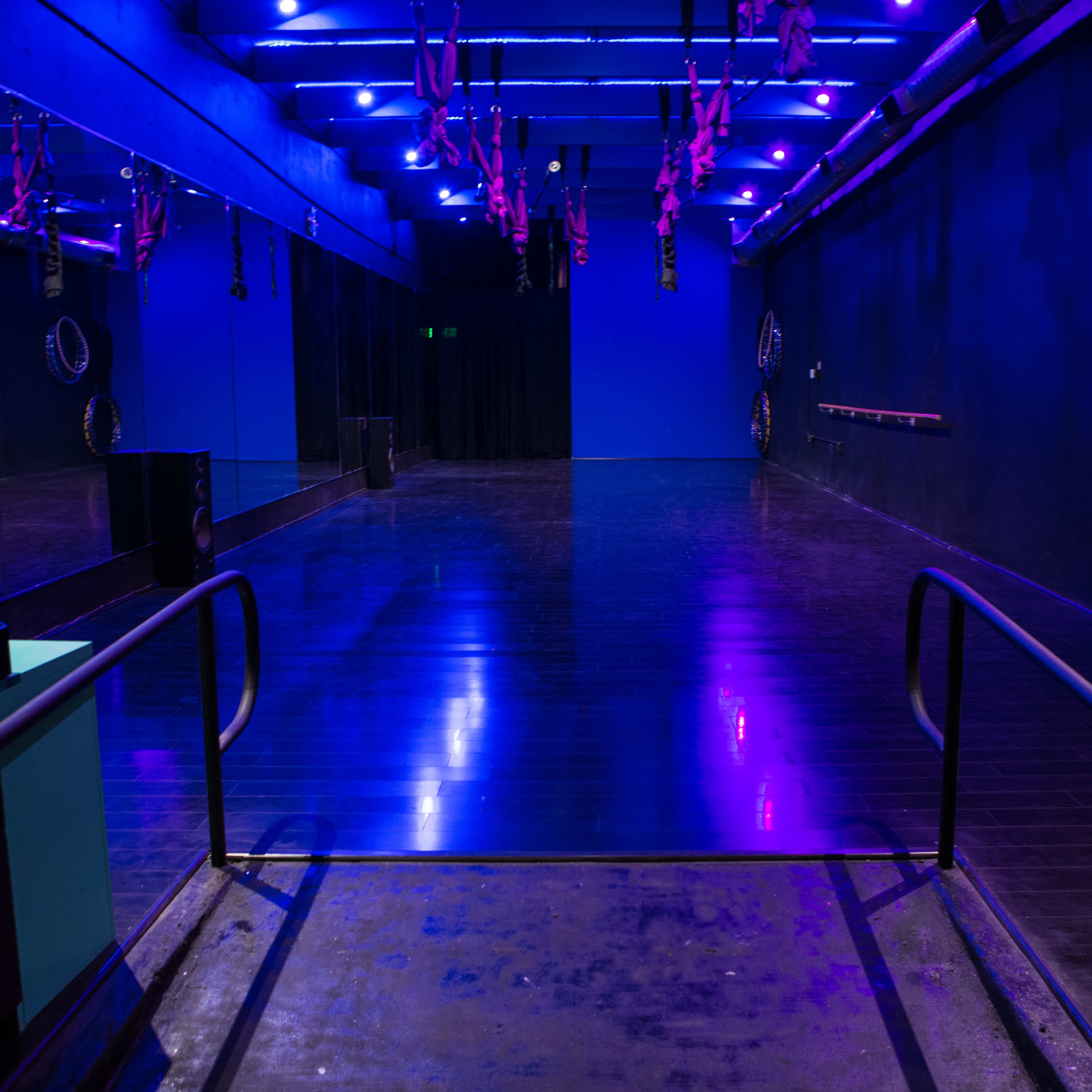 A spacious dance studio features a polished floor, vibrant blue walls, and overhead purple lighting.