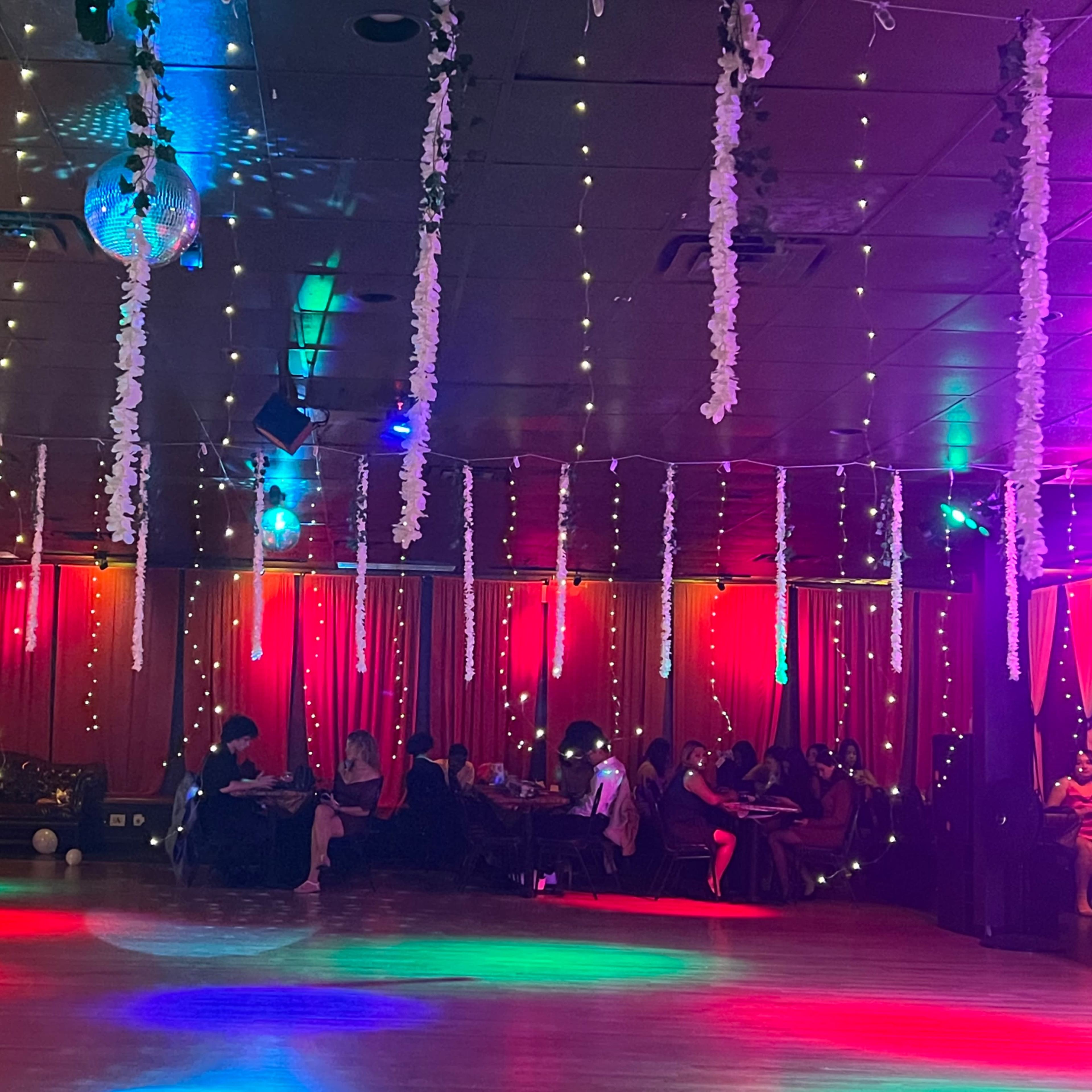 A decorated indoor venue features hanging lights and floral decorations, with people seated at tables in the background.
