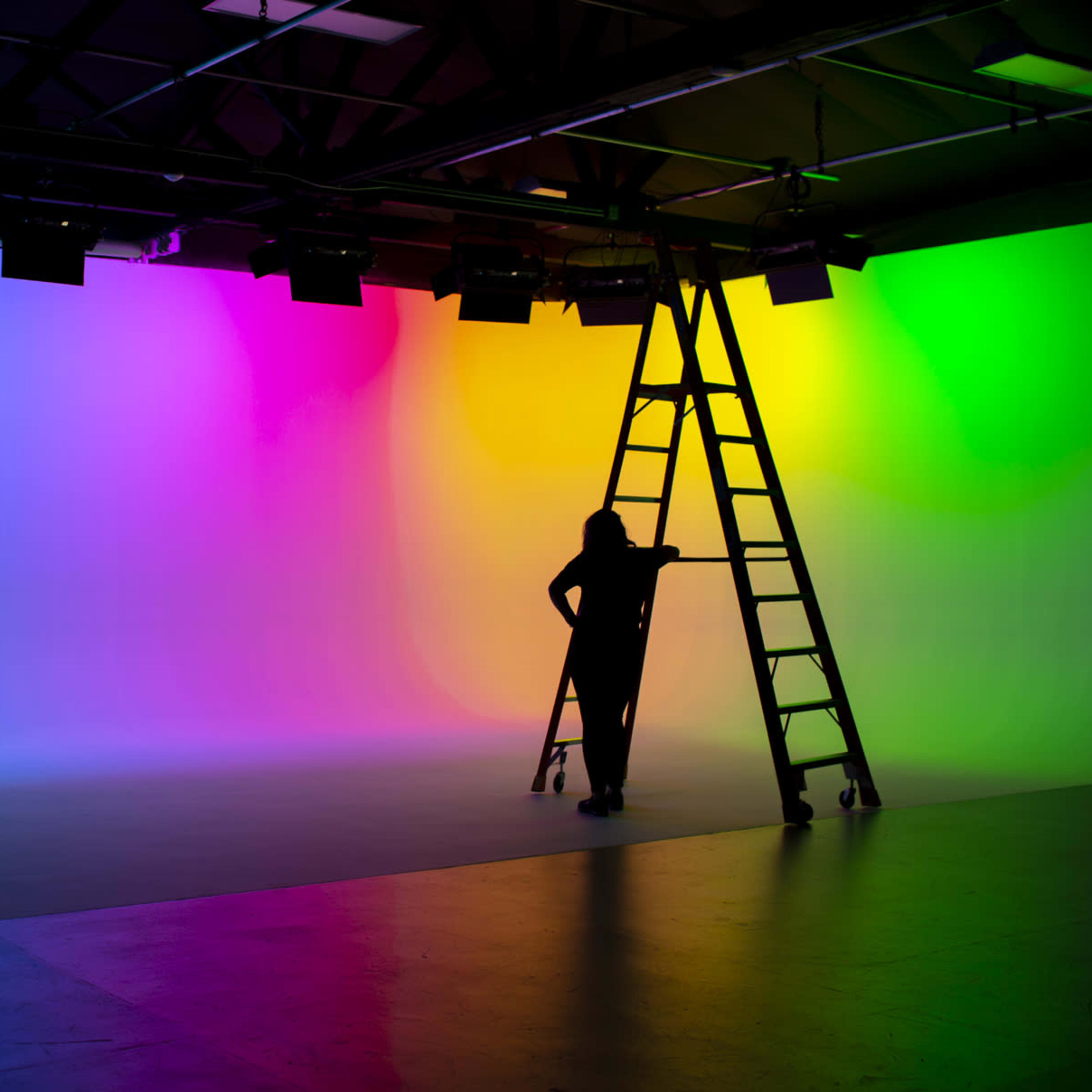 A person stands on a ladder in front of a brightly colored gradient backdrop in a studio.