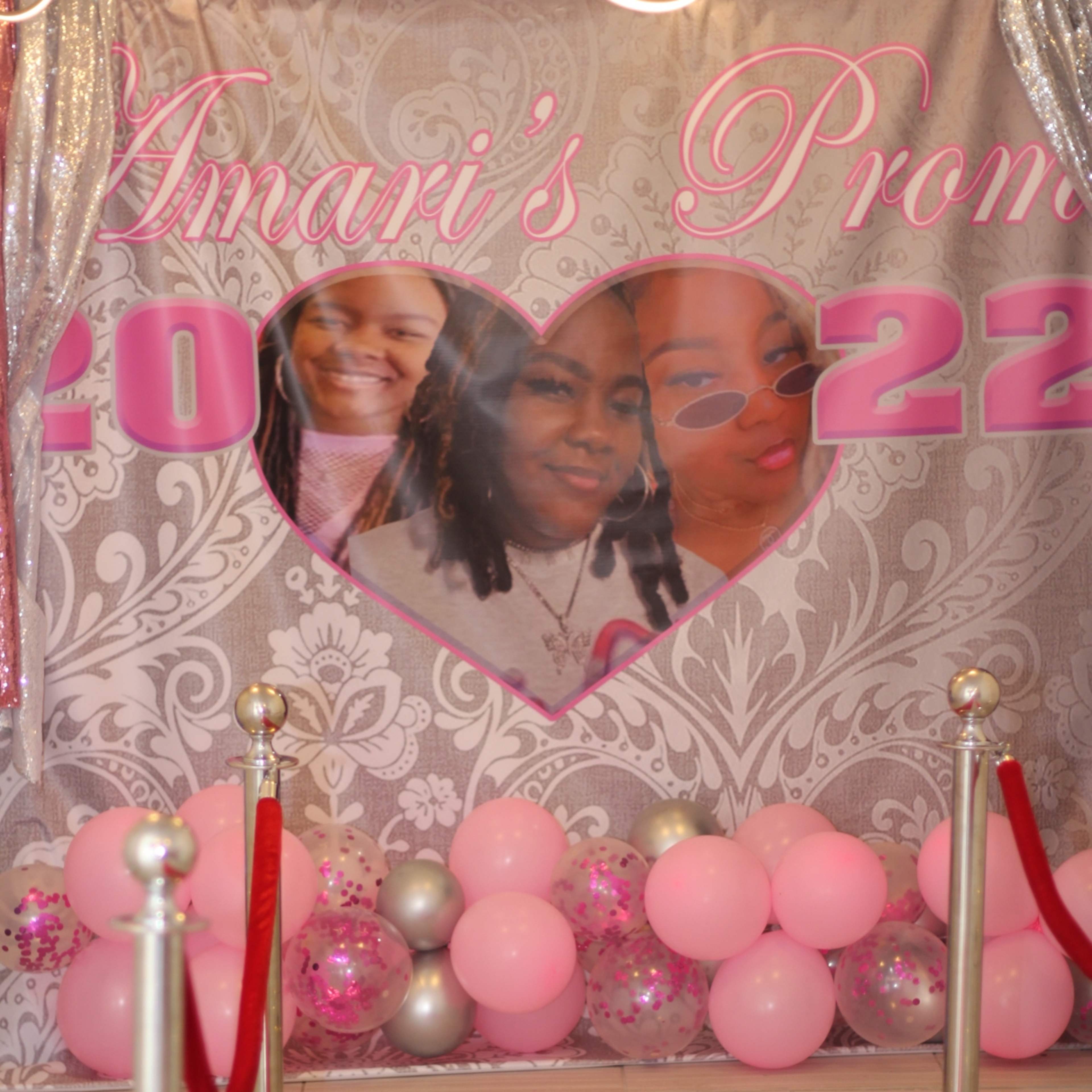 The scene features a decorated backdrop with images of three young women, a red carpet leading towards it, and pink balloon arrangements at the base.
