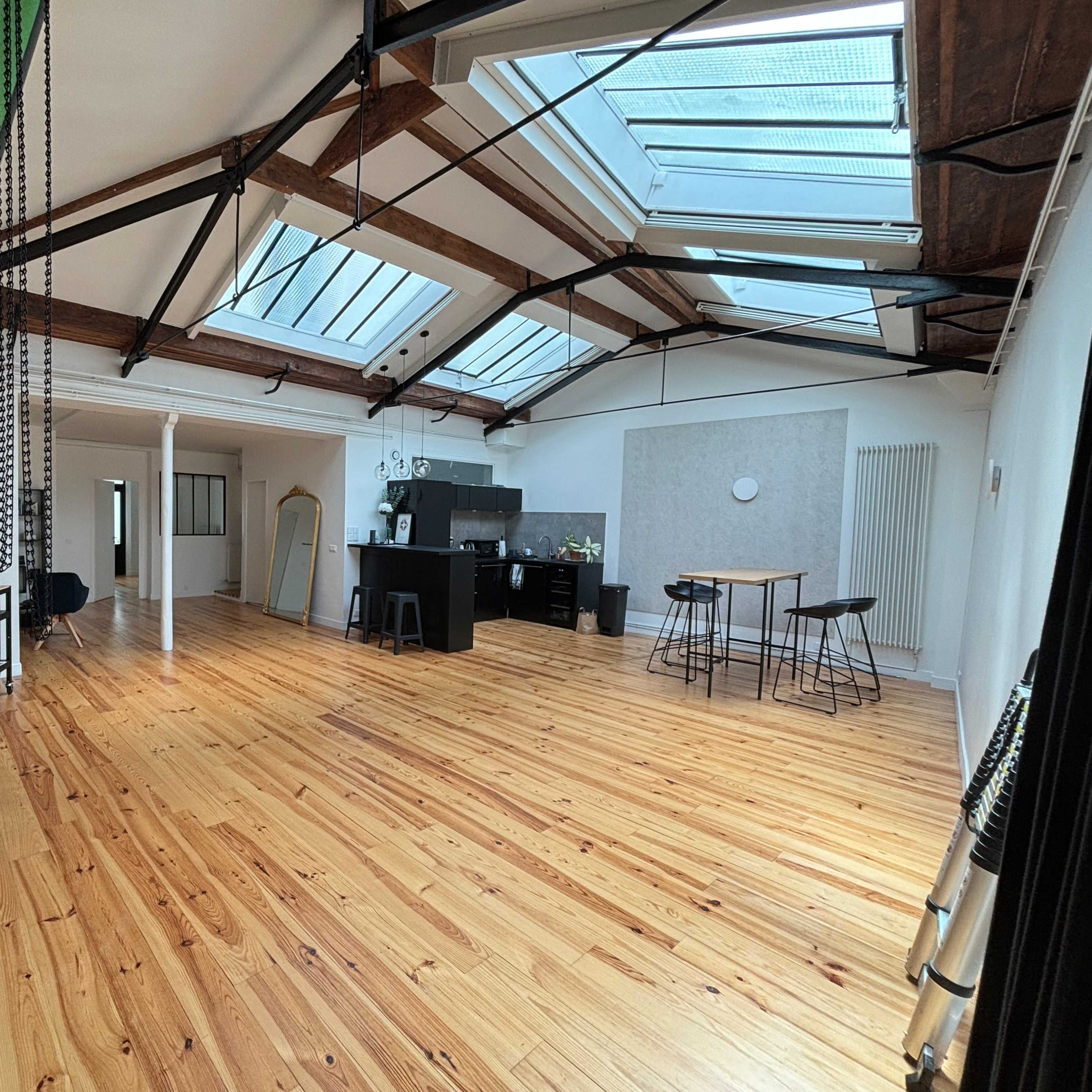 A spacious room with wooden flooring, large skylights, and various furniture arrangements including a kitchen area, tables, and chairs.