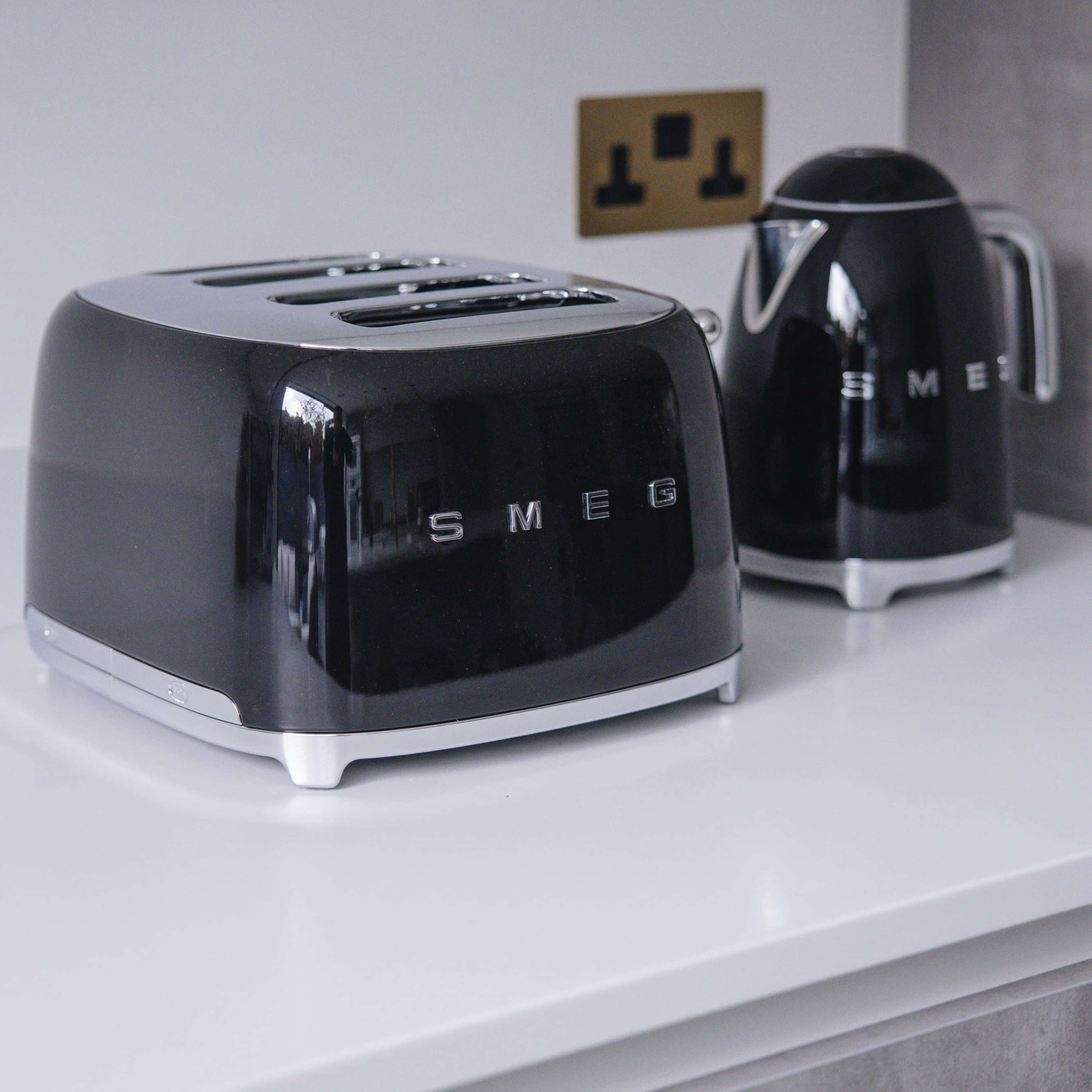 A black SMEG toaster and kettle are placed on a white countertop next to an electrical outlet.