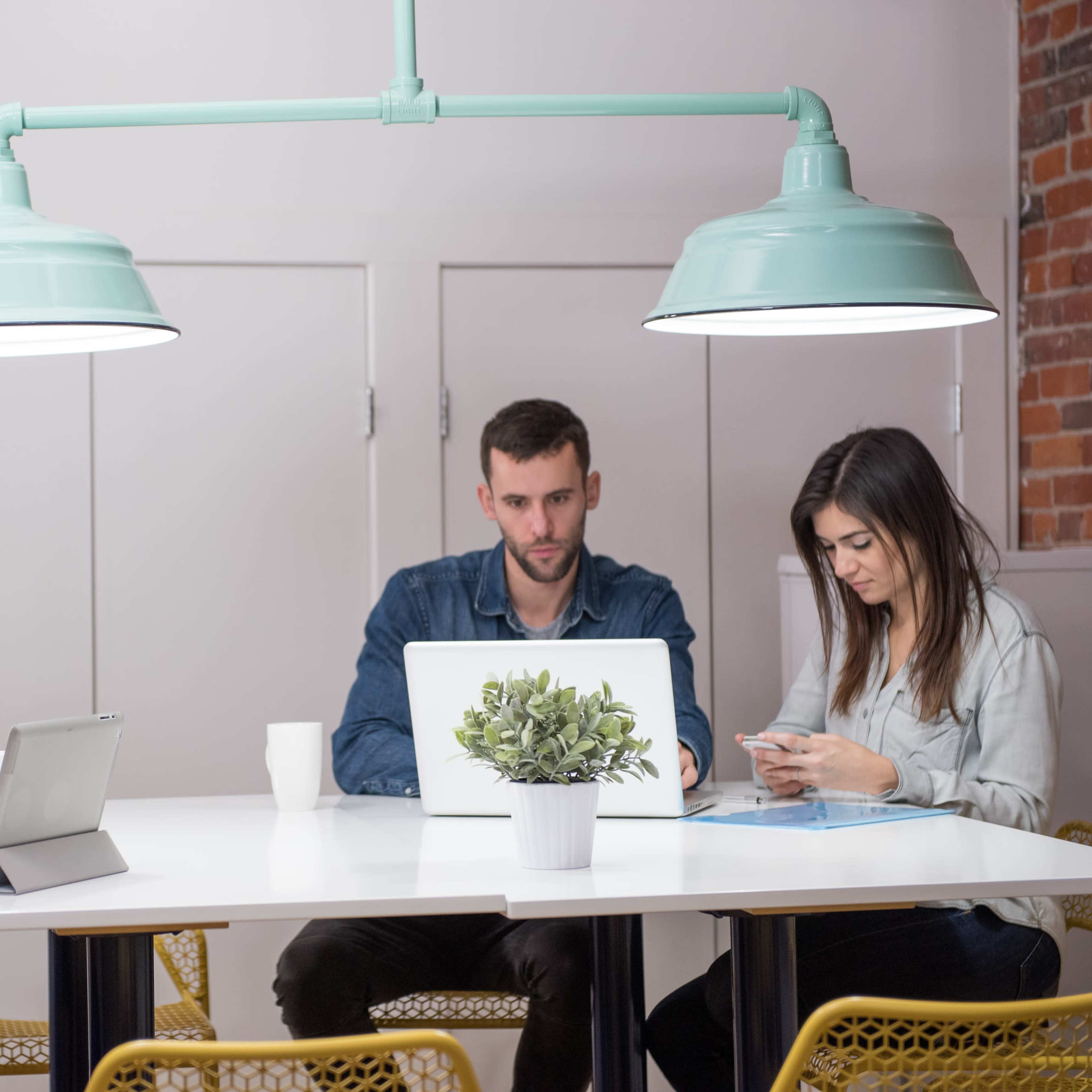 Three people are seated around a table in a modern office space, each using a device: one on a tablet, one on a laptop, and one on a smartphone.