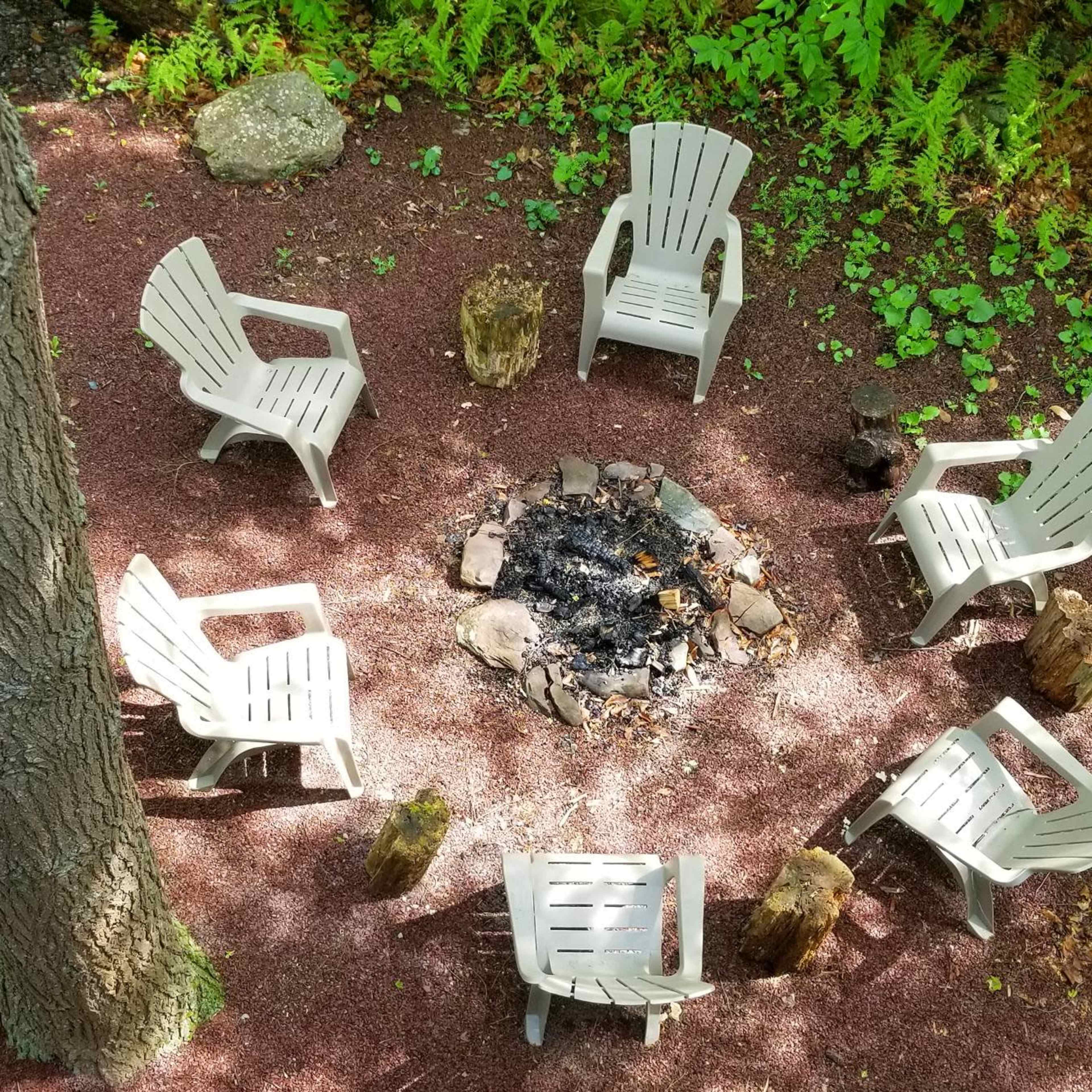 A circle of plastic chairs surrounds a fire pit in a forested area.