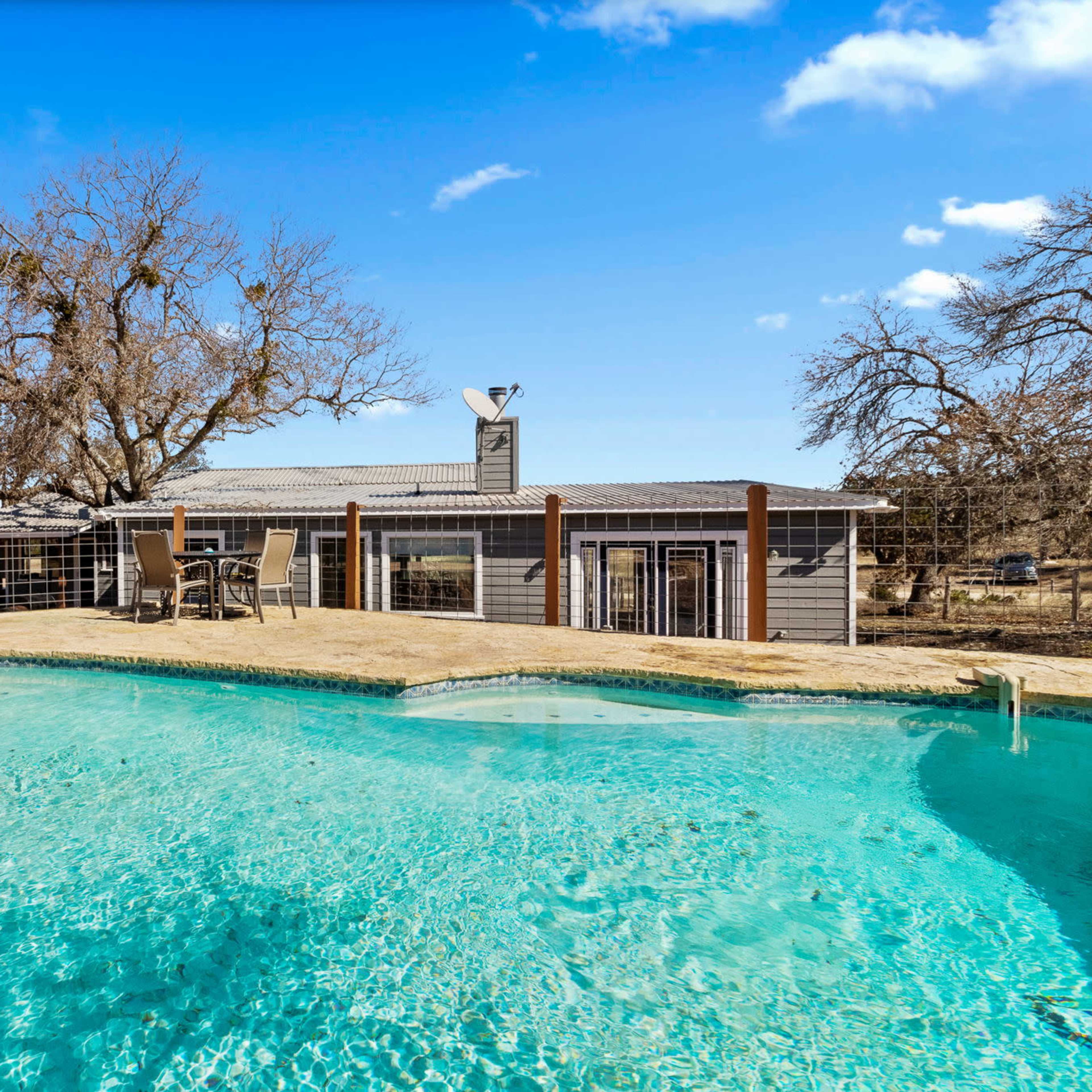 A clear swimming pool in the foreground with a modern house and bare trees in the background under a blue sky.