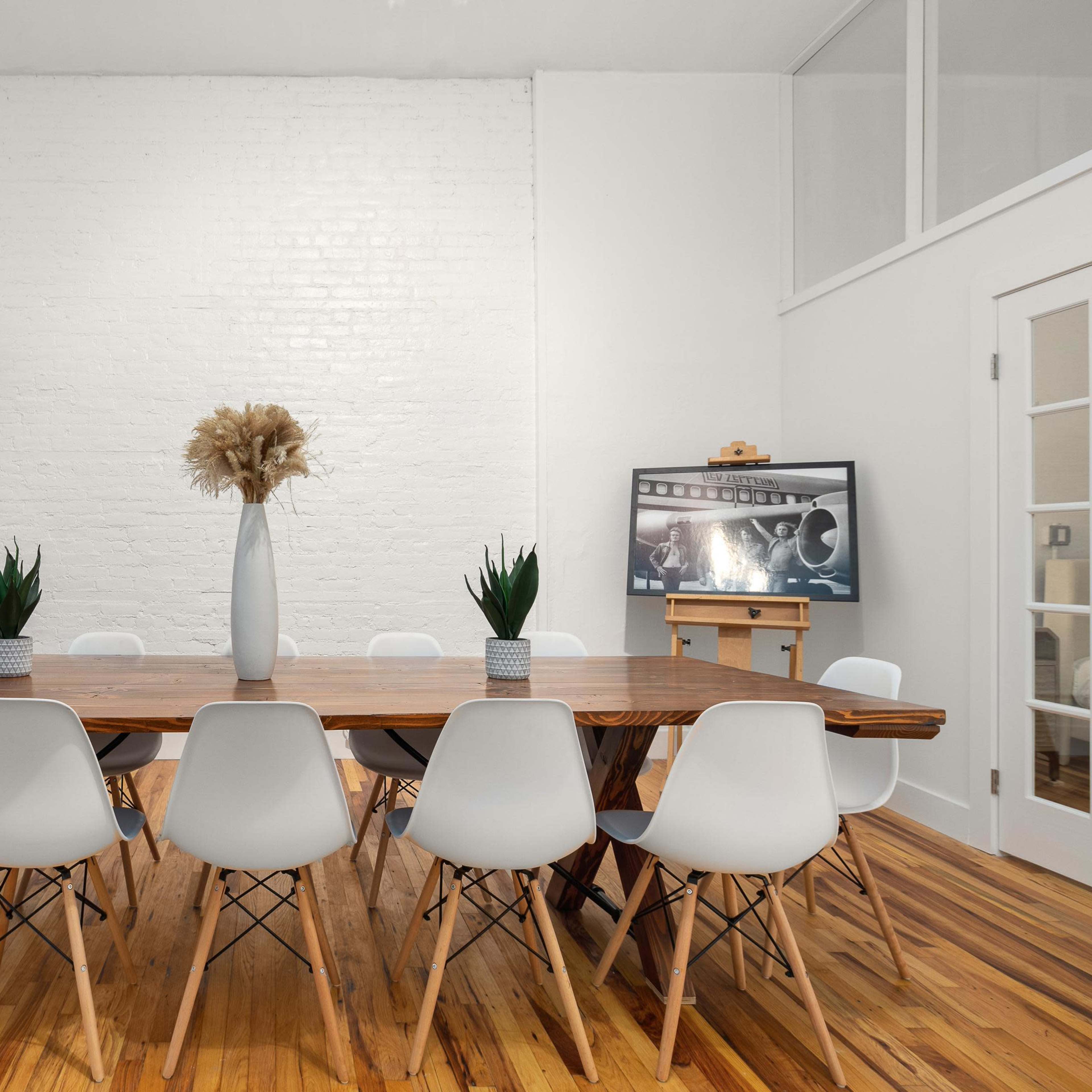 A spacious meeting room features a long wooden table surrounded by white chairs, with potted plants, a large vase, and a screen displaying images on one side.