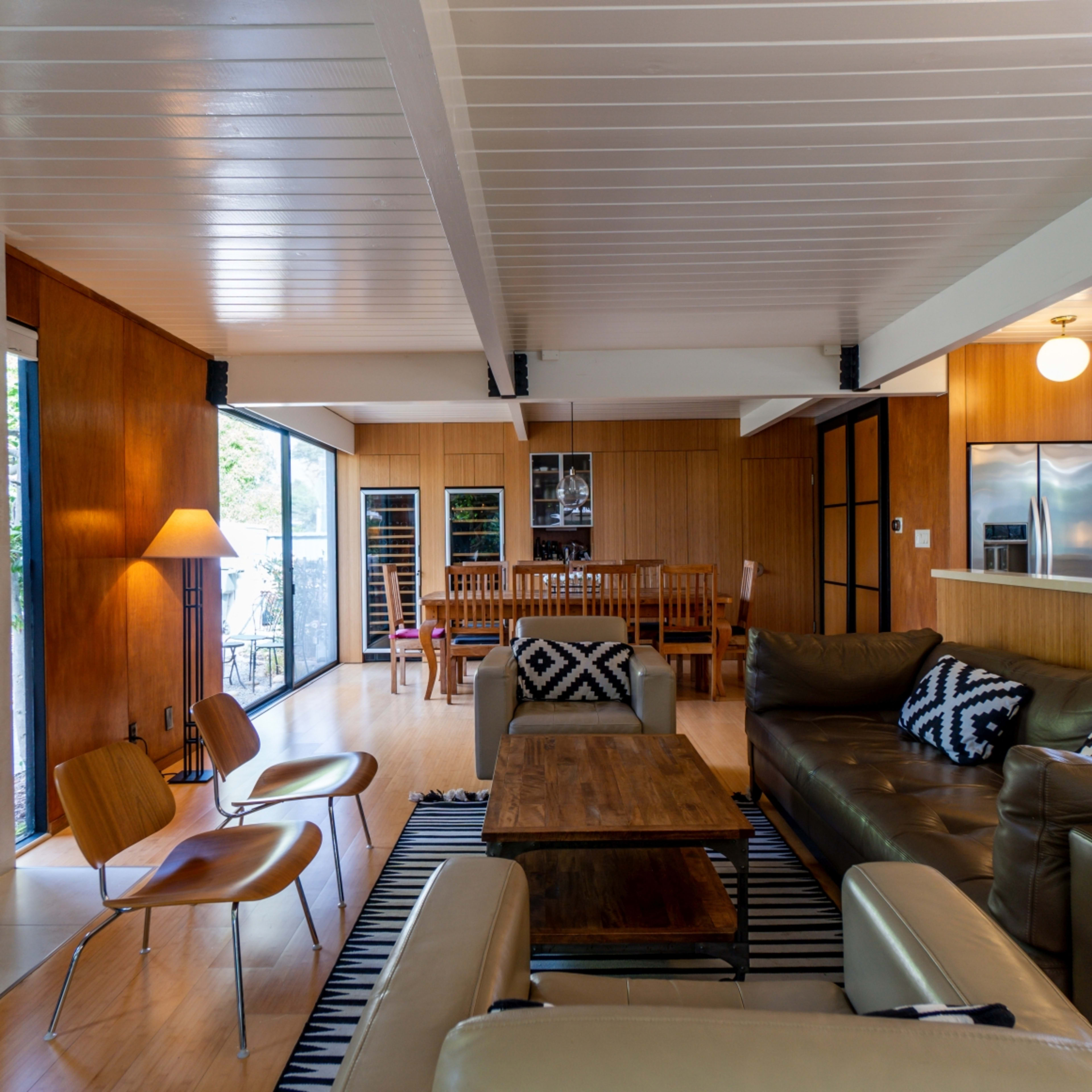 A modern interior living space with a brown leather sectional sofa, a wooden coffee table, a dining area in the background, and large windows overlooking an outdoor patio.