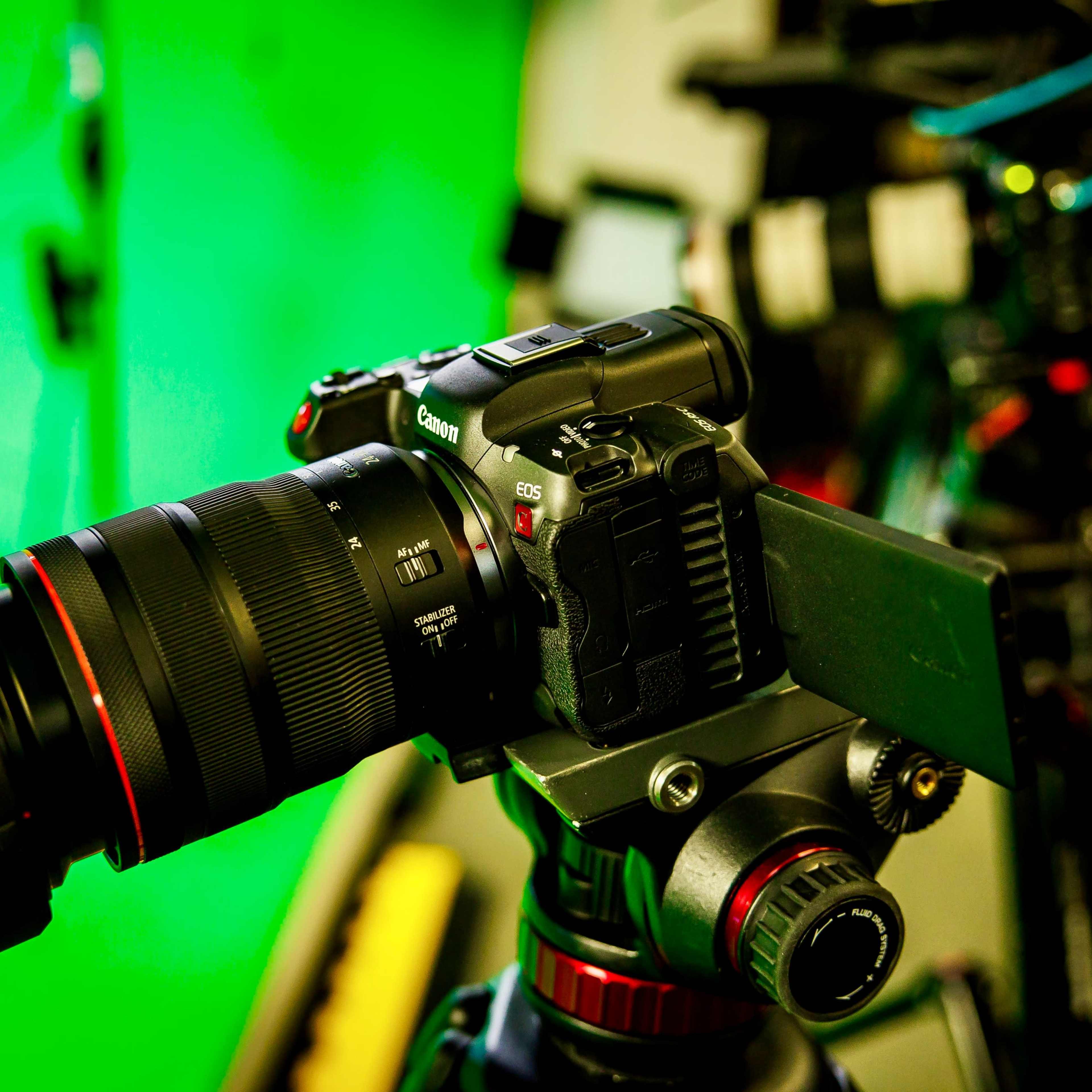 A Canon camera is mounted on a tripod in front of a green screen backdrop.