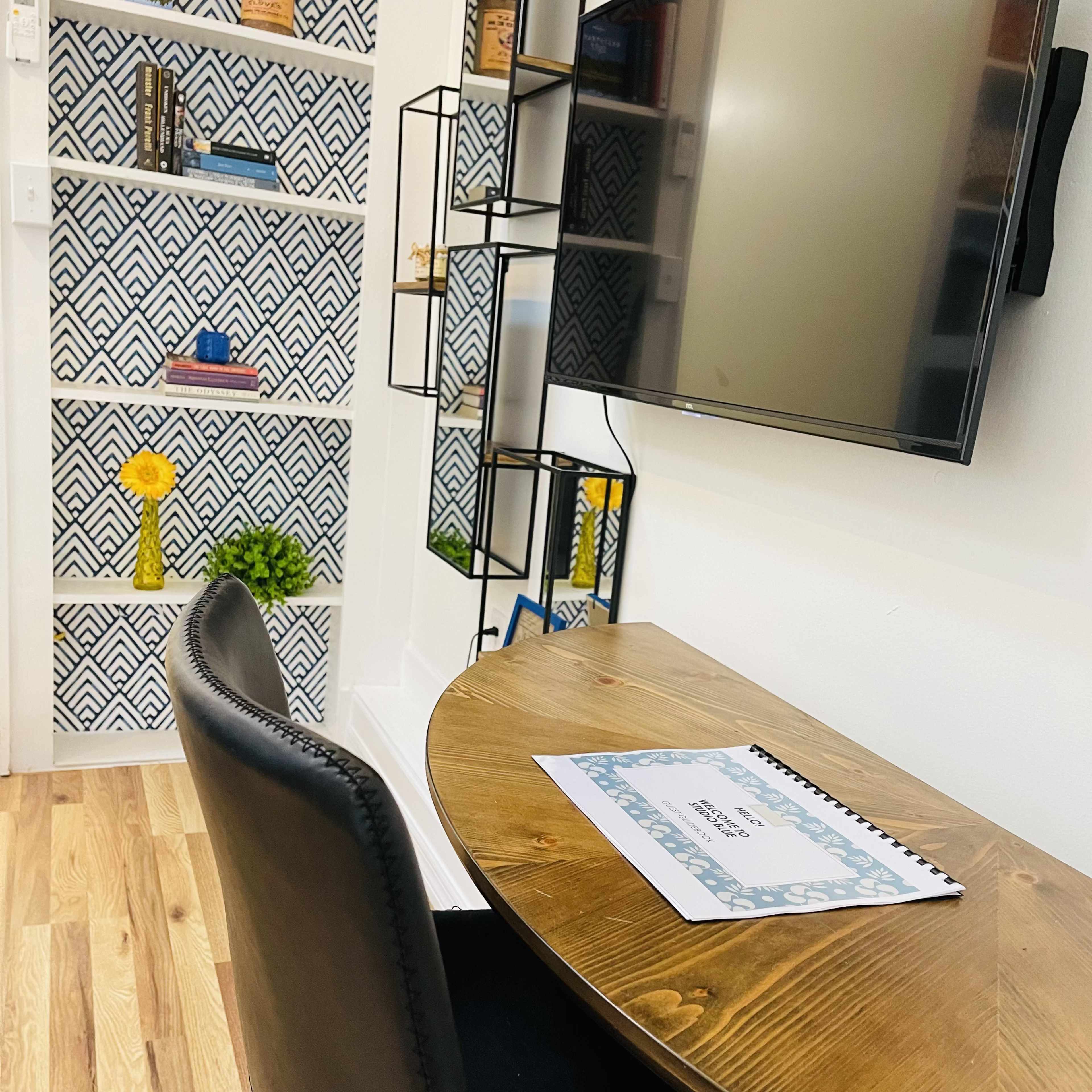 A small workspace with a round wooden table, a black chair, and a wall-mounted TV, featuring patterned shelving in the background.
