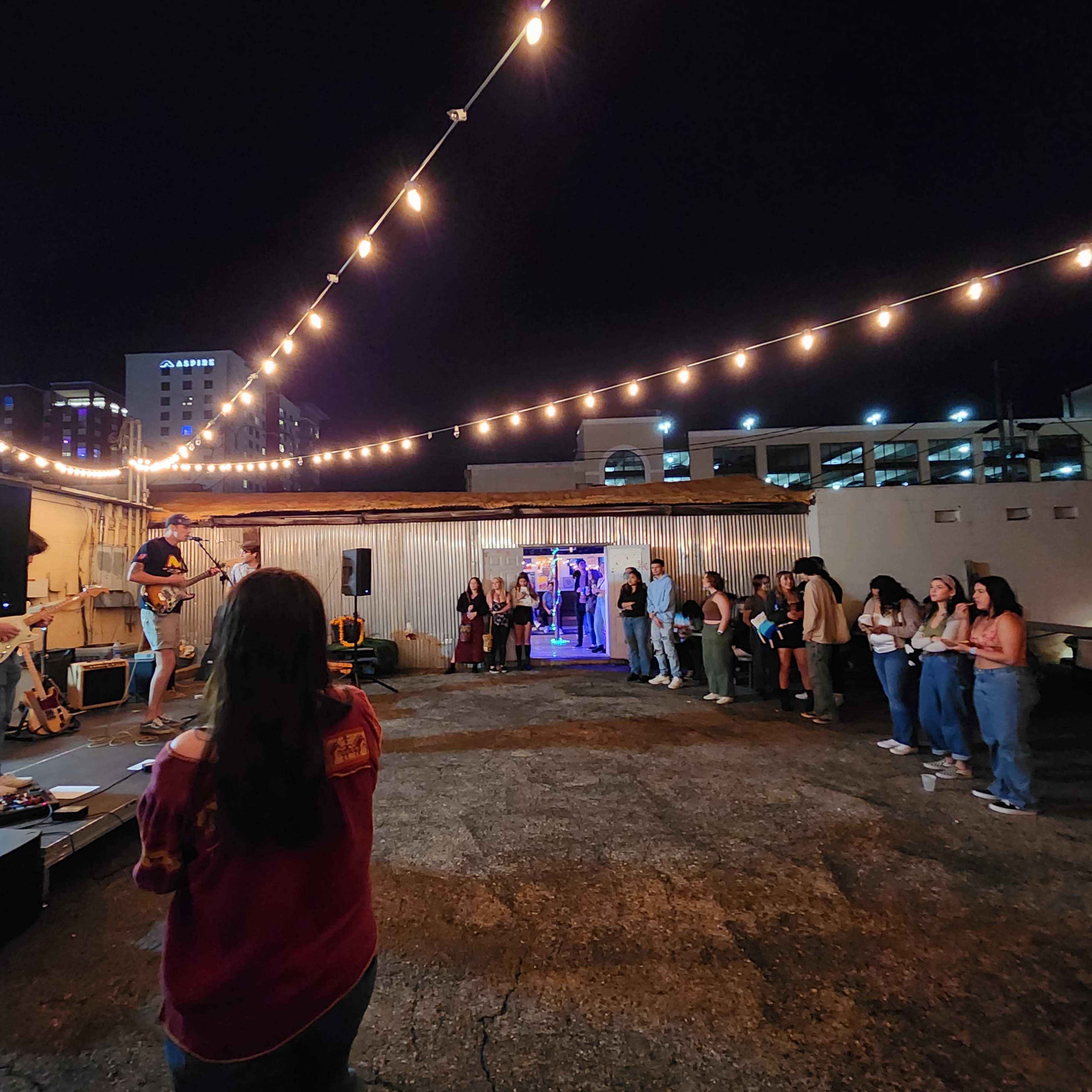 A musician performs on a rooftop stage in front of a small crowd under string lights at night.