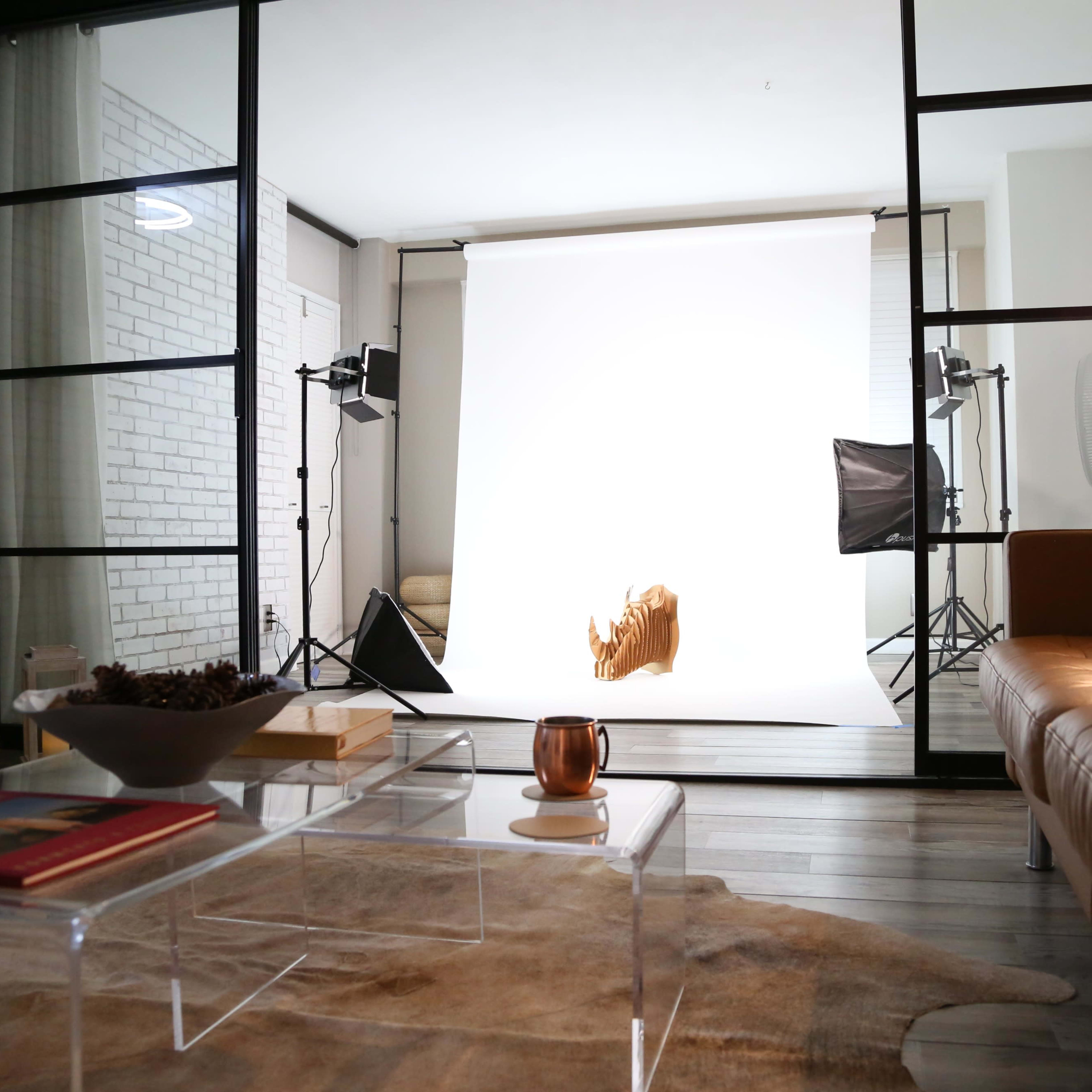 A photography studio setup with a white backdrop, softbox lights, and a cozy living area visible in the foreground.