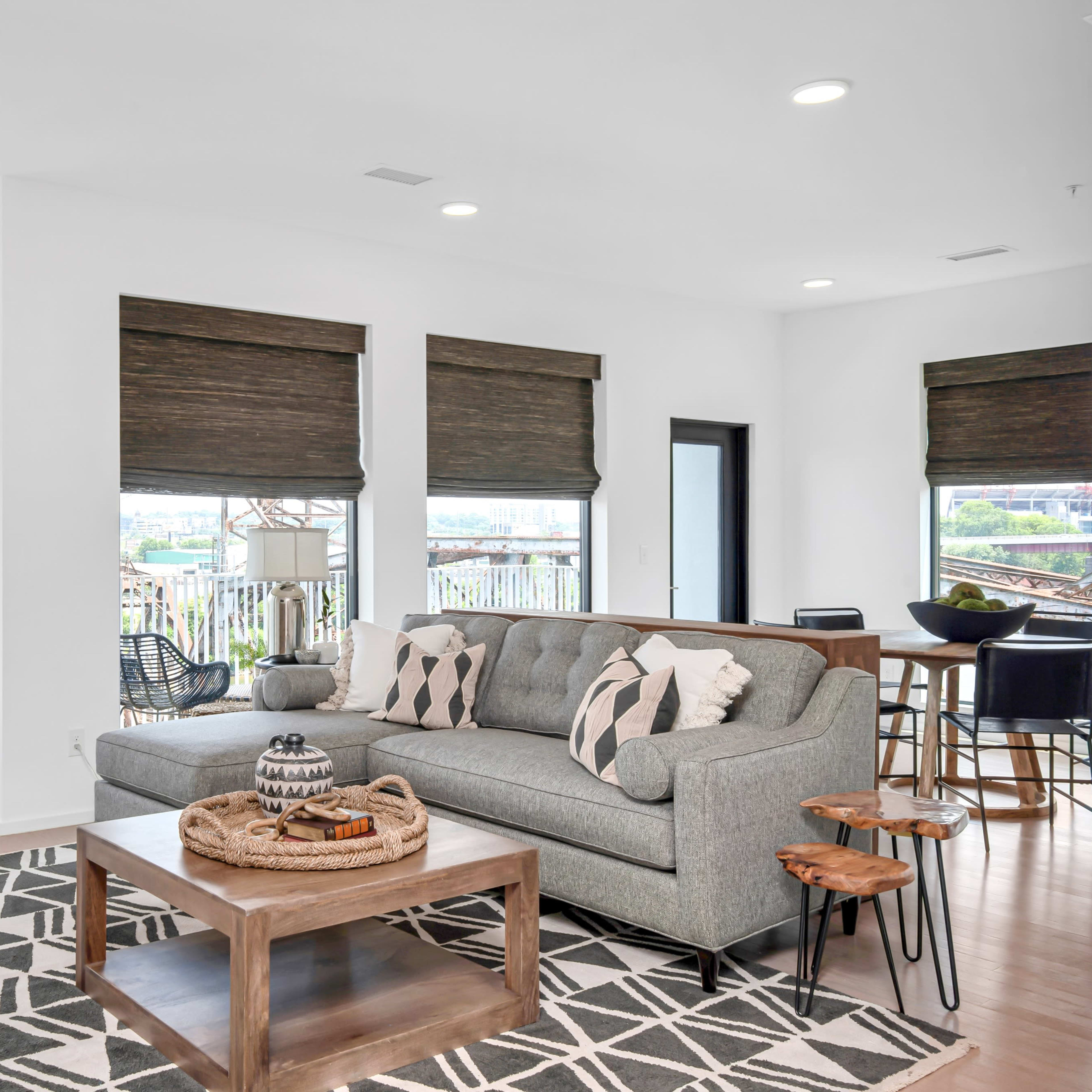 A modern living room features a gray sofa, wooden coffee table, and patterned rug, with large windows showing a view of the outdoors.
