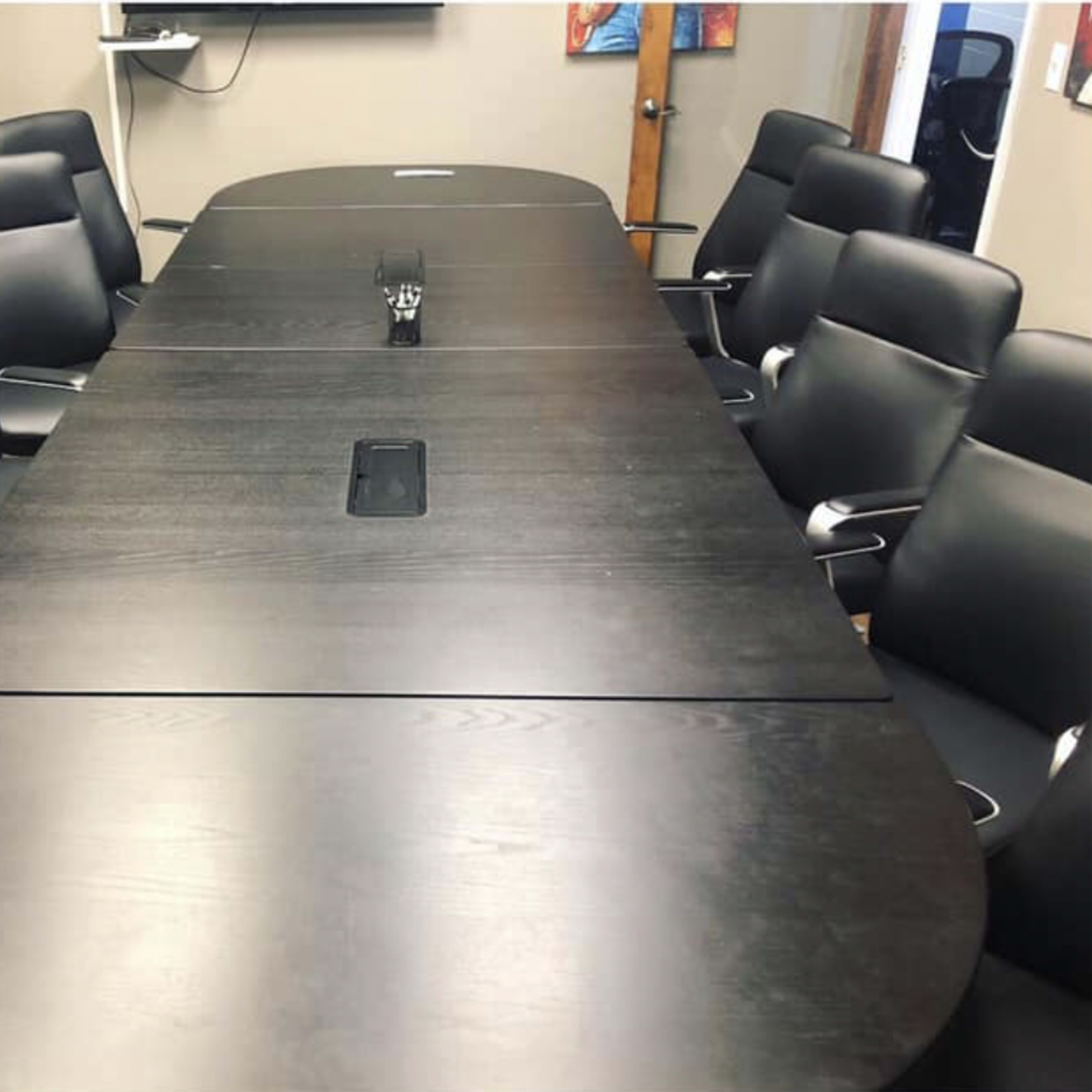 A large oval conference table surrounded by black ergonomic chairs is set up in a meeting room.