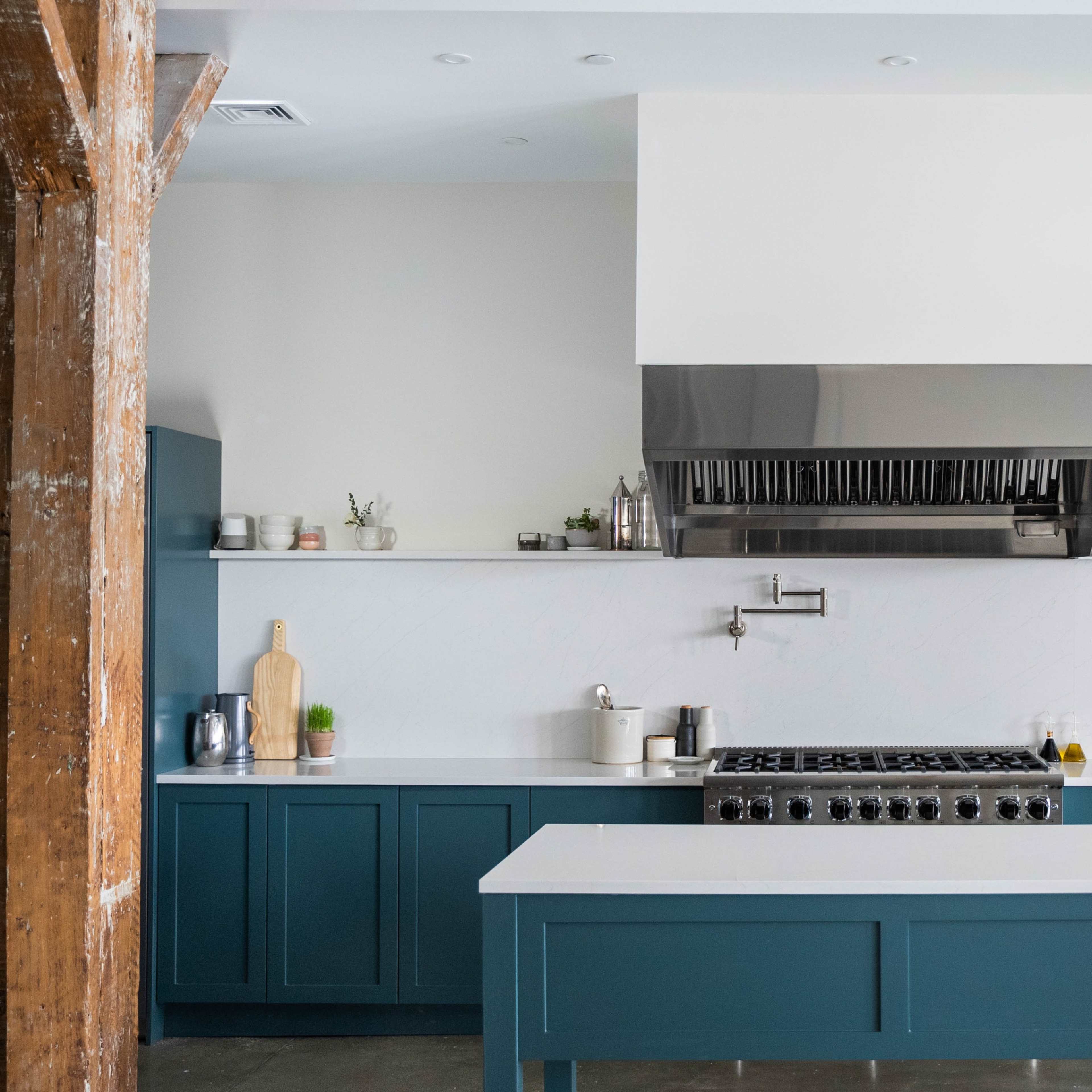 A modern kitchen features a large island with a stove, stainless steel hood, and minimalist cabinetry in teal and white.