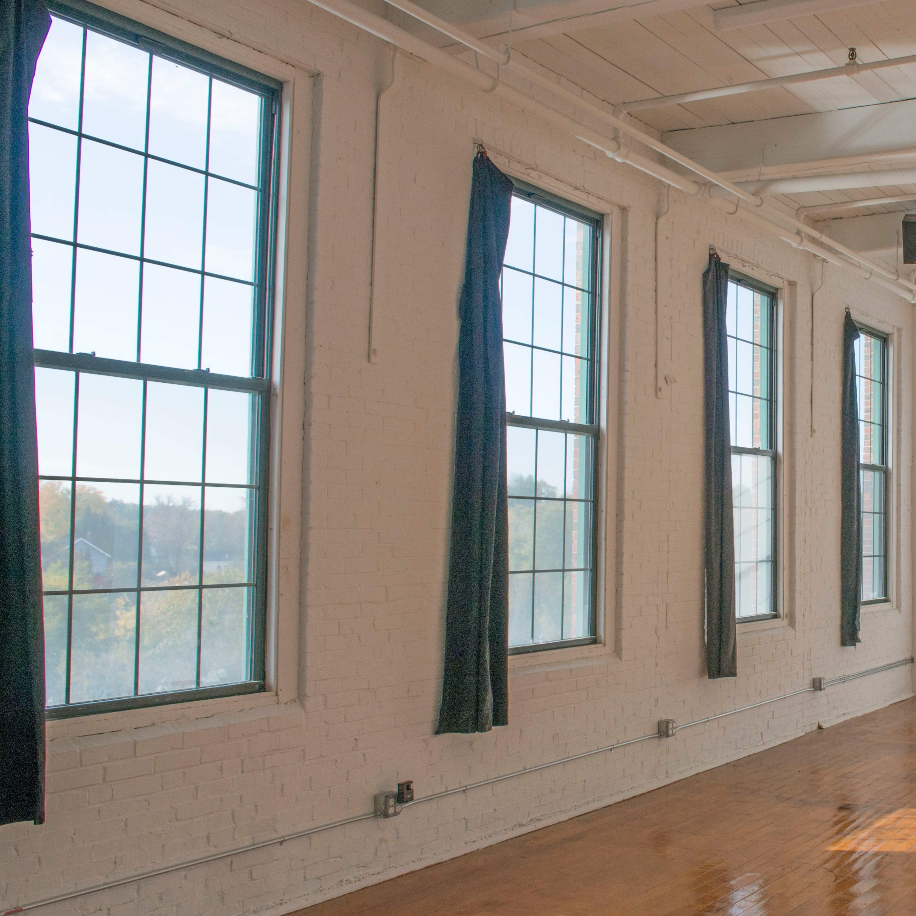 A bright, empty room features large windows and wooden flooring, with light casting shadows on the wall.