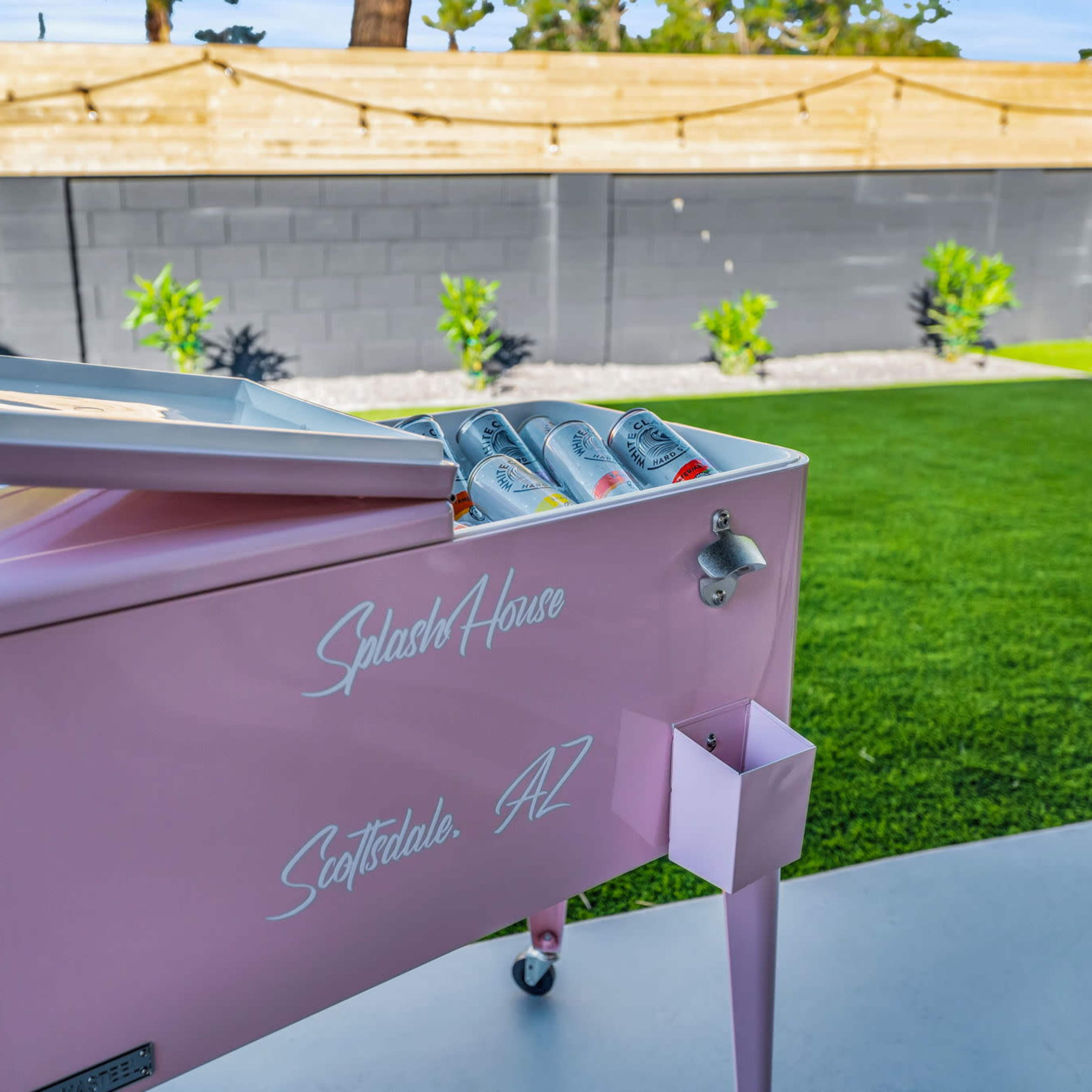 A pink cooler sits on a patio, open and displaying beverages, with a green lawn and a wooden fence in the background.