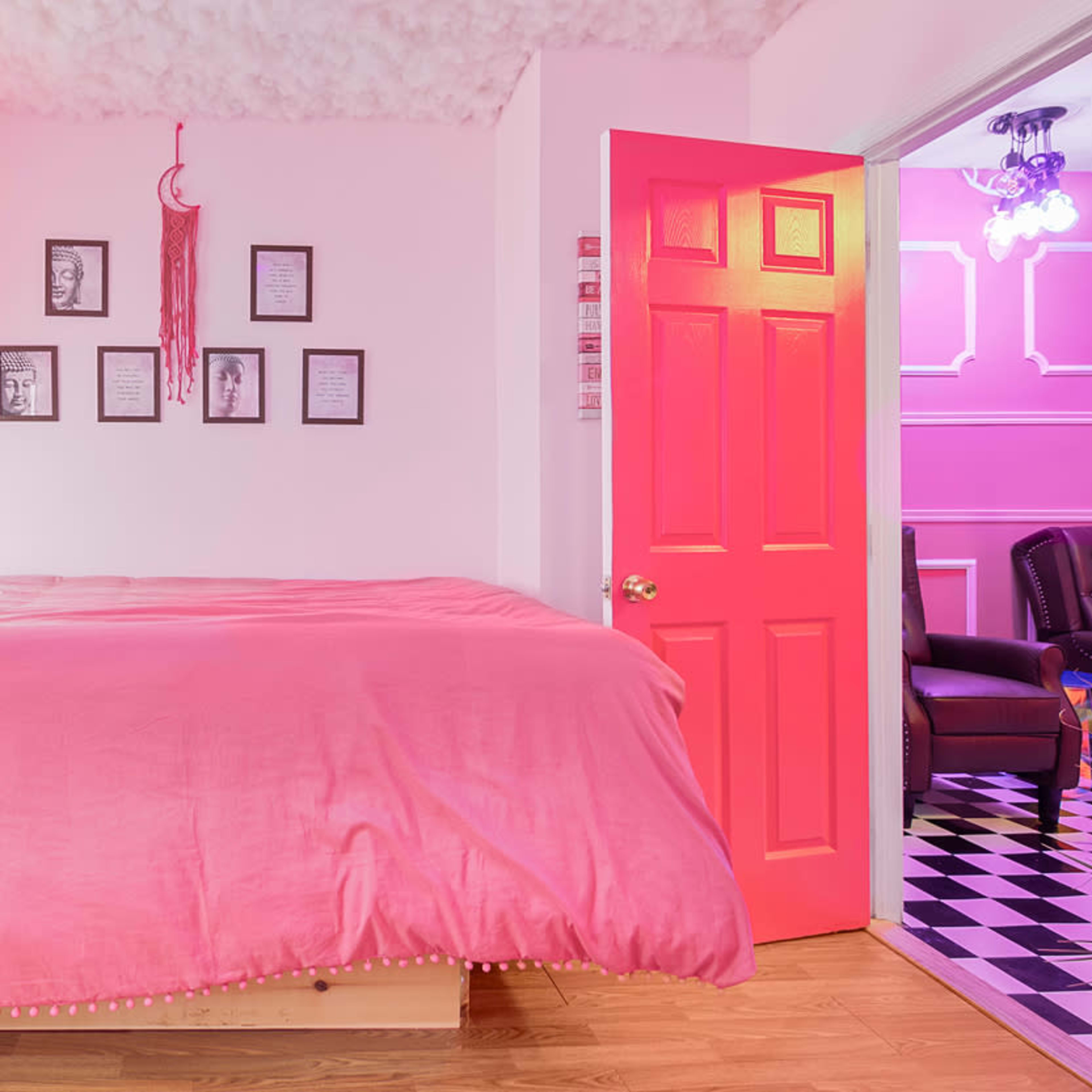 A bright pink bedroom features a large bed with a matching bedspread and framed art on the walls, along with a vibrant pink door leading to another room.