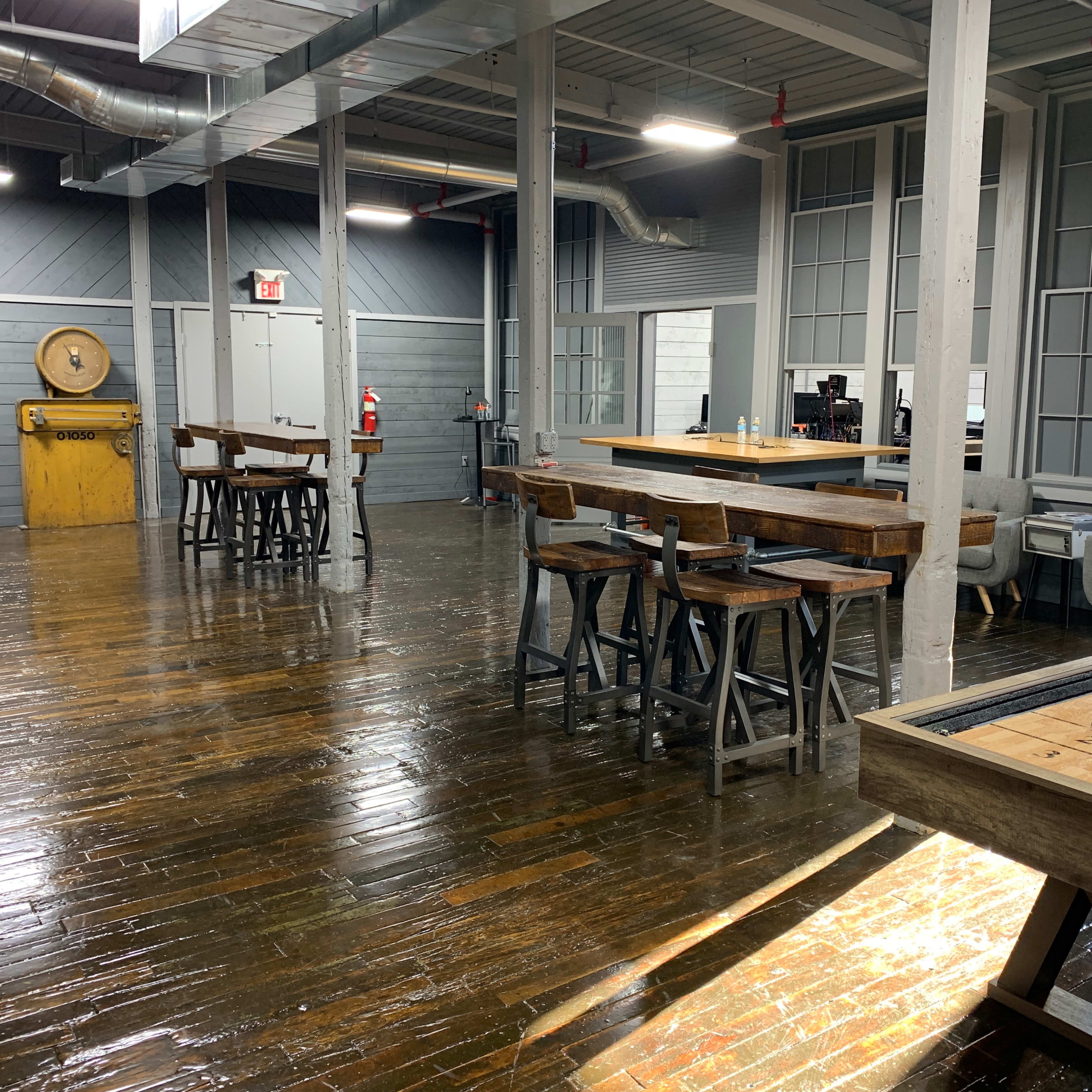 A spacious interior with polished wood floors, several wooden tables and chairs, a bar counter, and large windows allowing natural light.