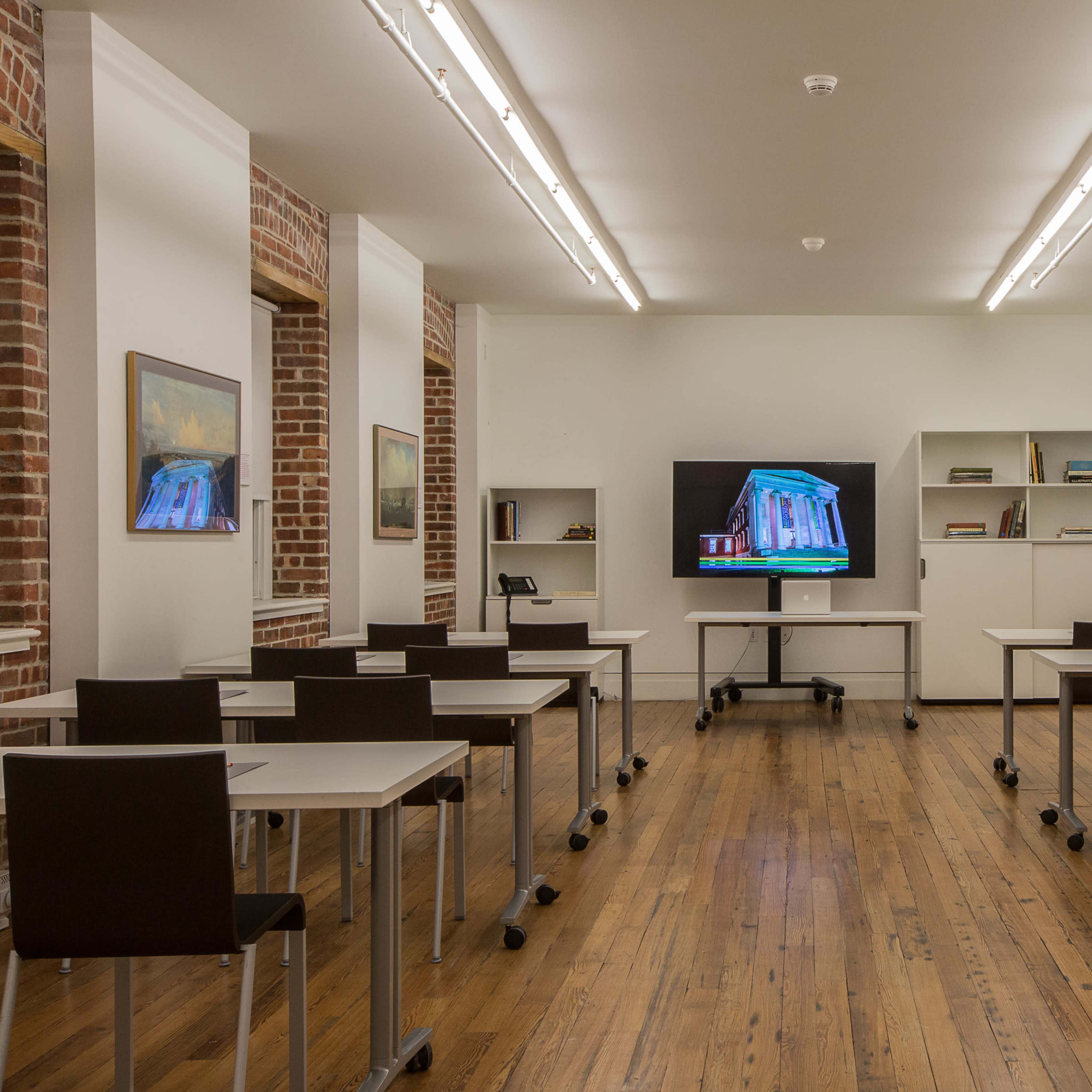 A well-lit classroom with rows of modern desks and a large screen at the front, surrounded by brick walls and shelves.