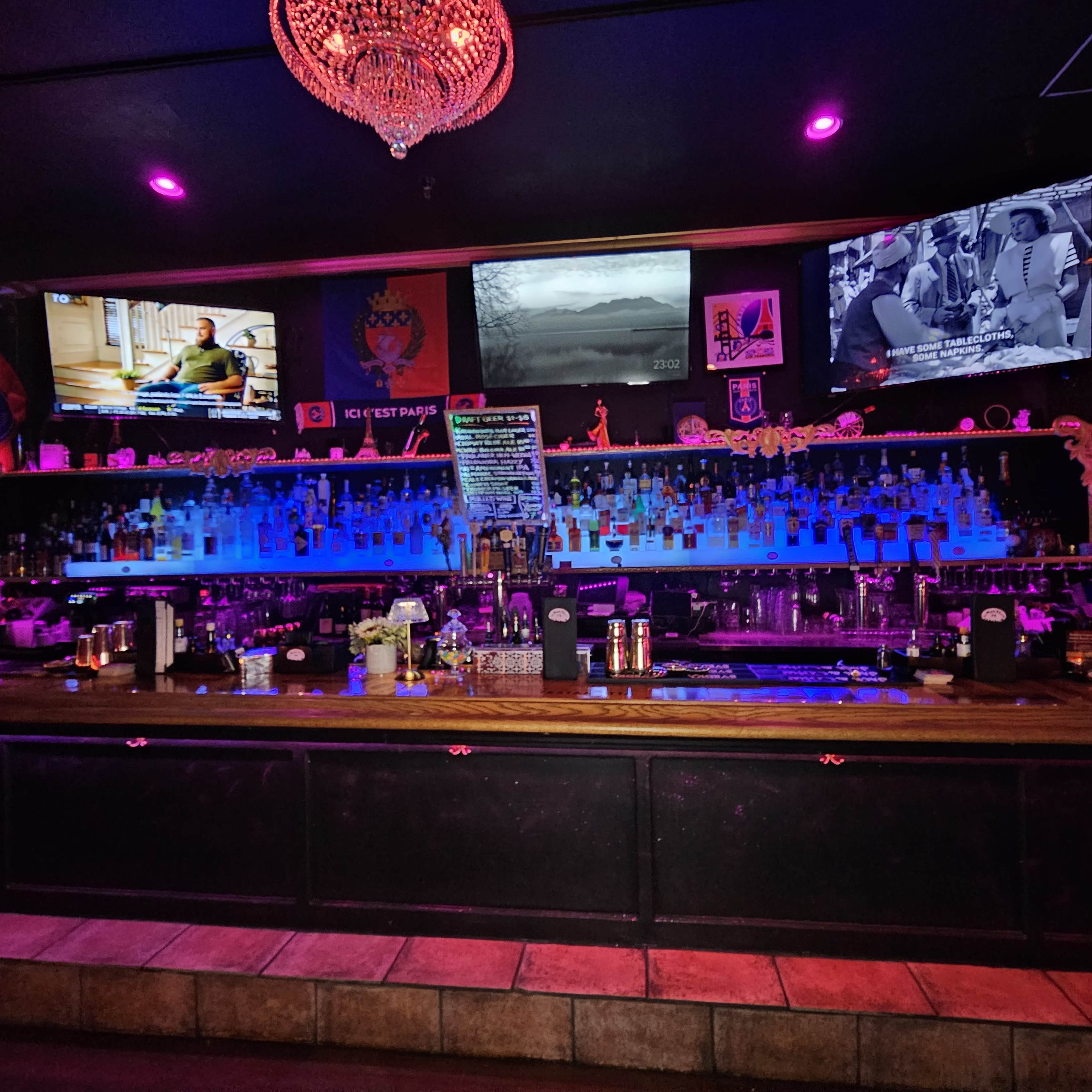 A dimly lit bar with a long counter featuring various bottles of alcohol and illuminated displays on the walls.