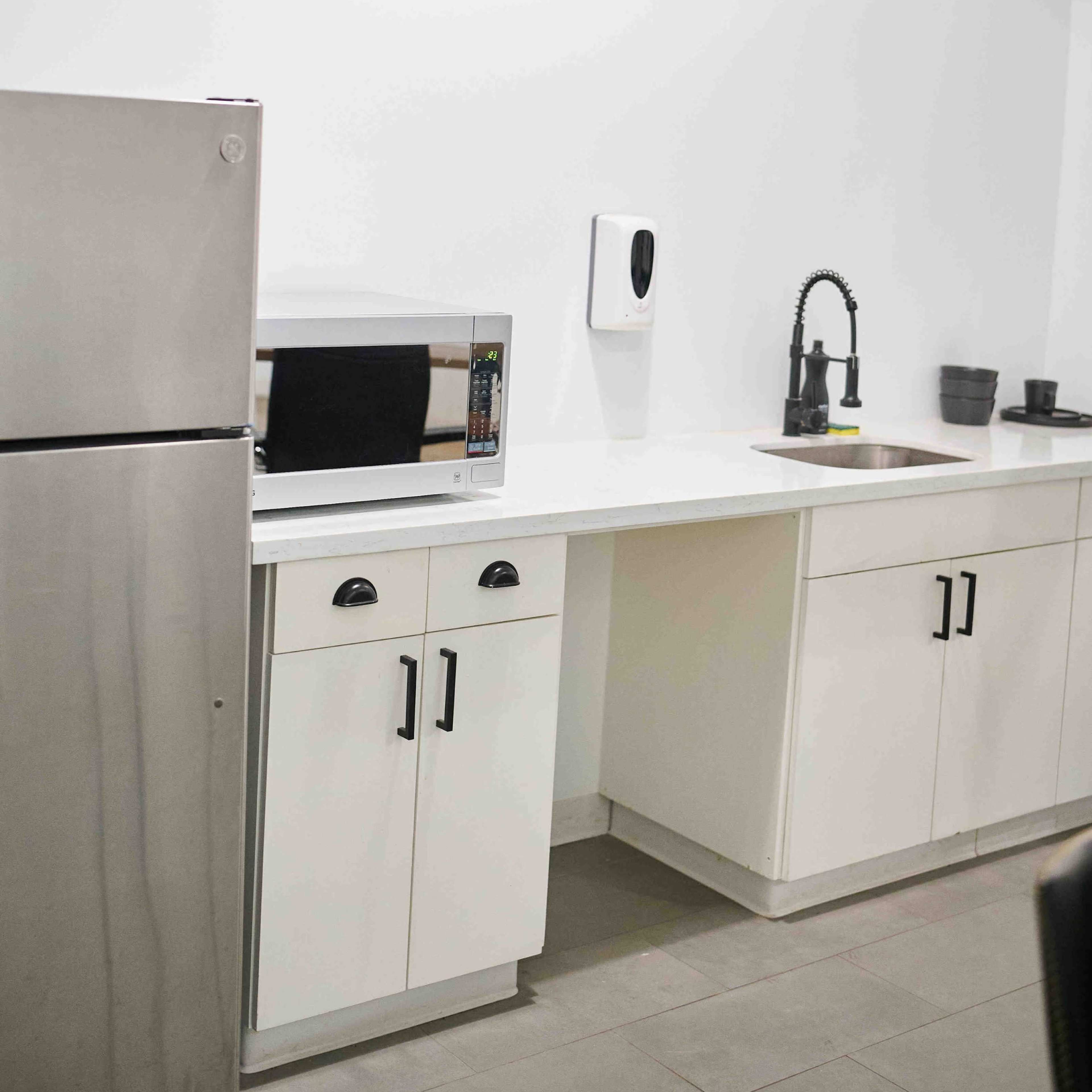 A modern kitchen with a stainless steel refrigerator, a microwave, a sink with a black faucet, and white cabinets against a light-colored wall.