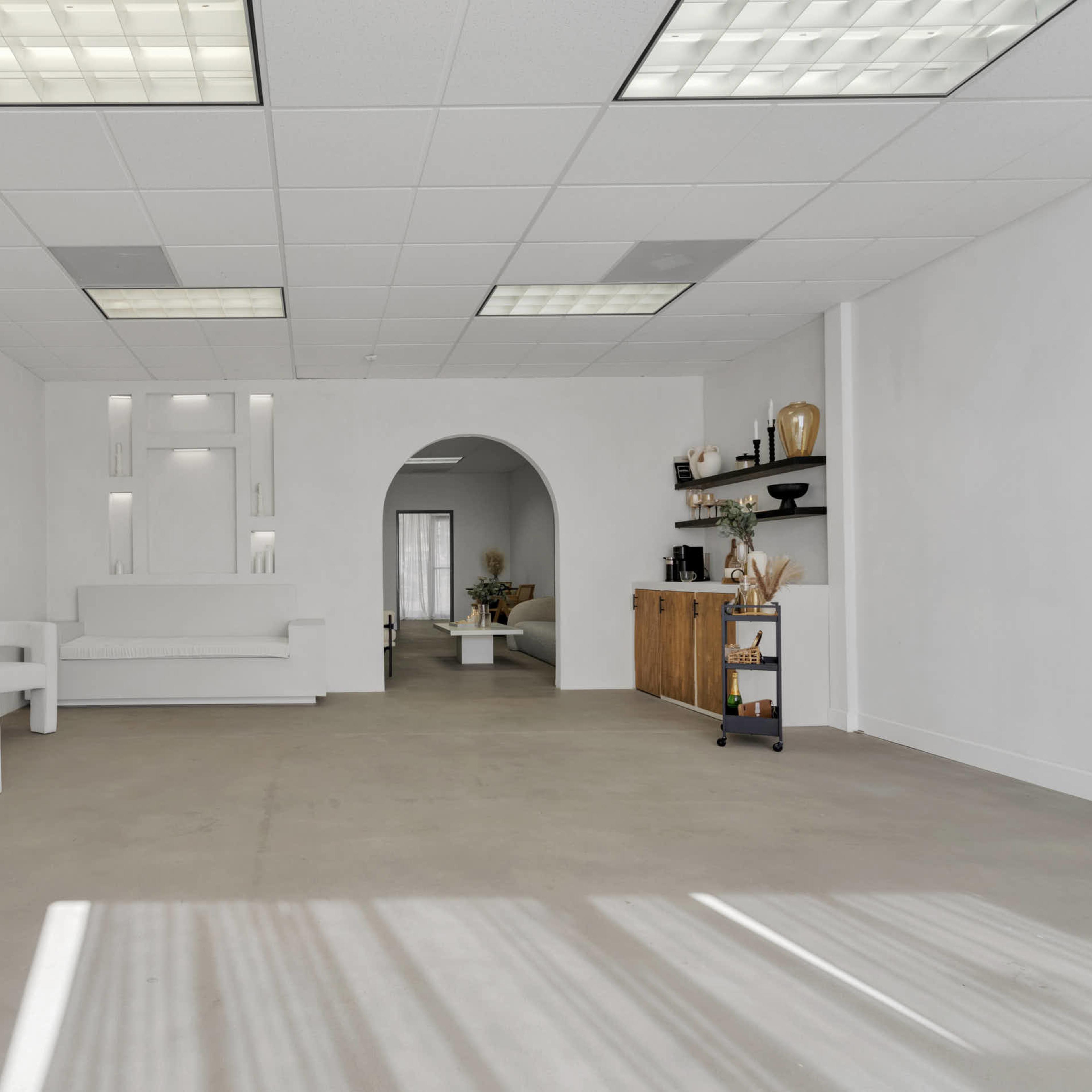 A minimalistic, white interior space featuring a raised platform, an archway, and simple furniture arrangements.