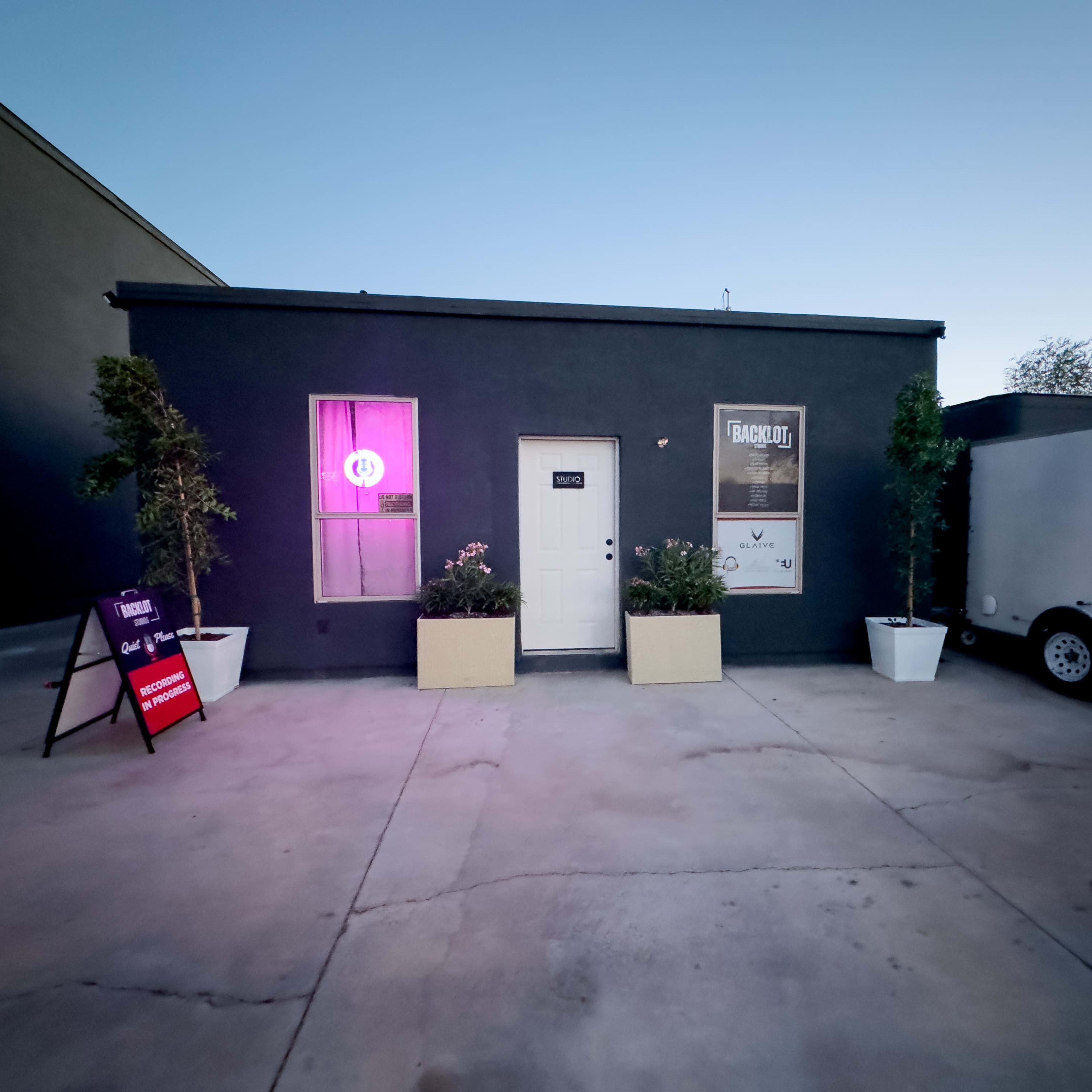A small gray building with a white door, flanked by potted plants, and illuminated by purple lighting from a window.